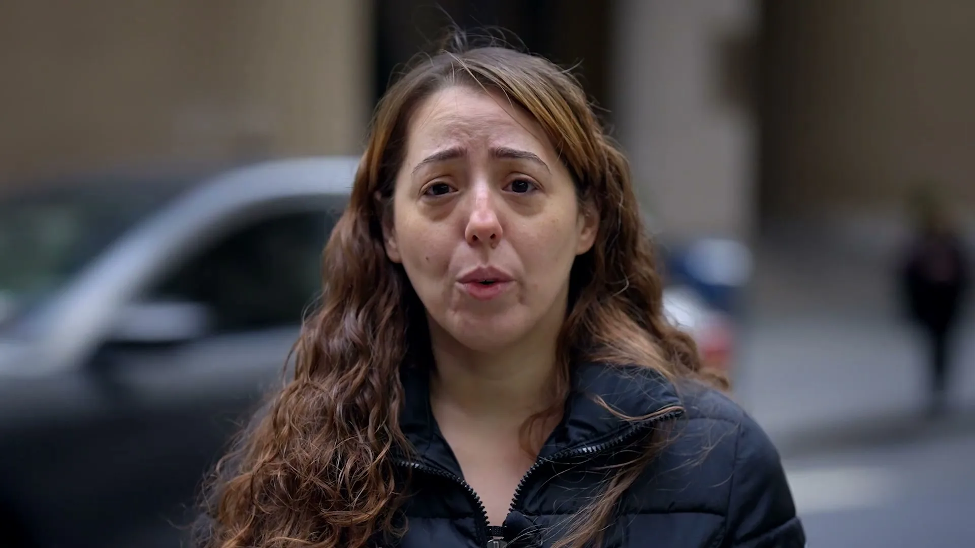 A young woman with long brown hair and a black puffer jacket speaks directly to the camera on a city street.