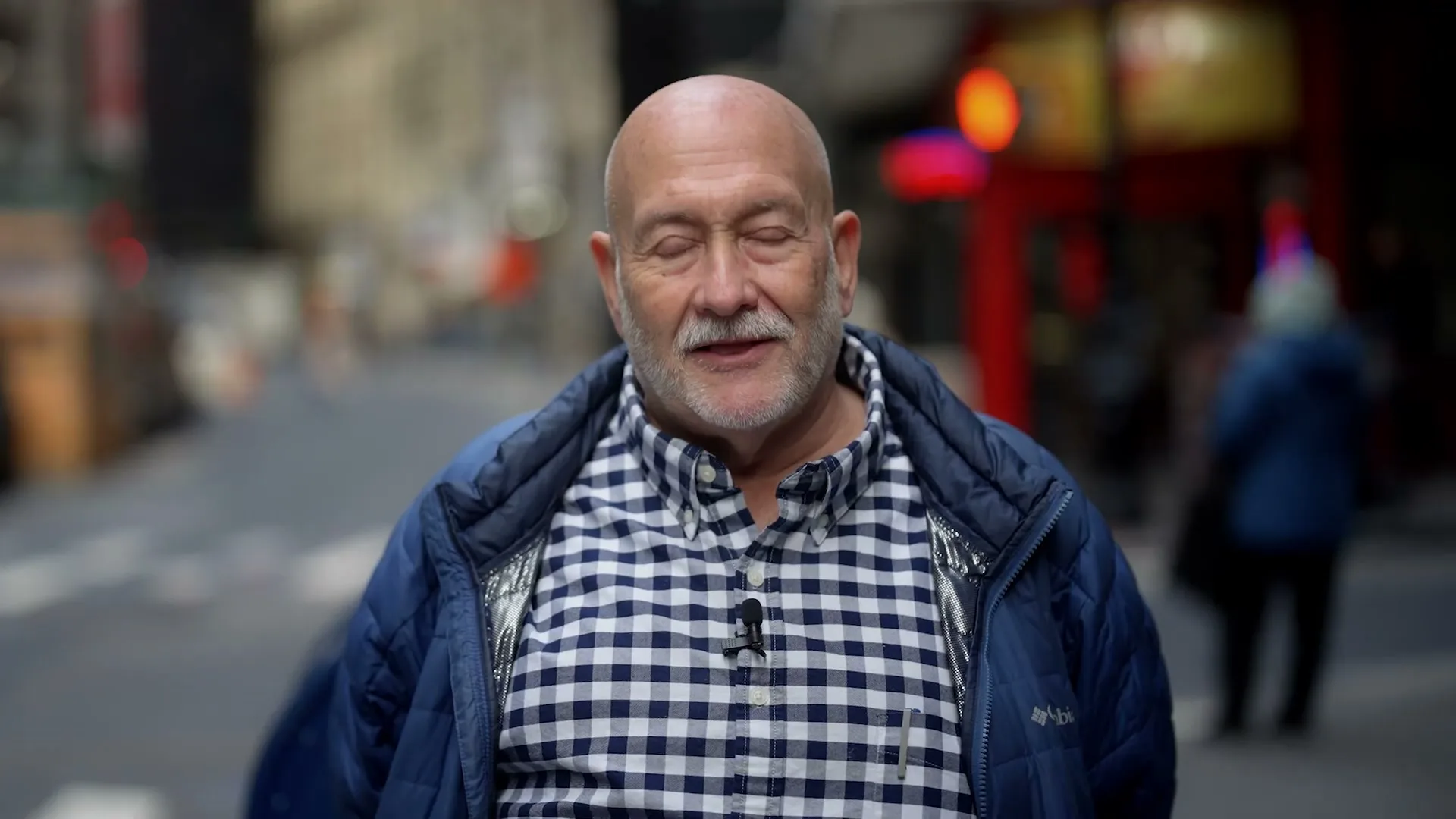 A bald, older man with a white beard and blue plaid shirt speaks directly to the camera on a city street.