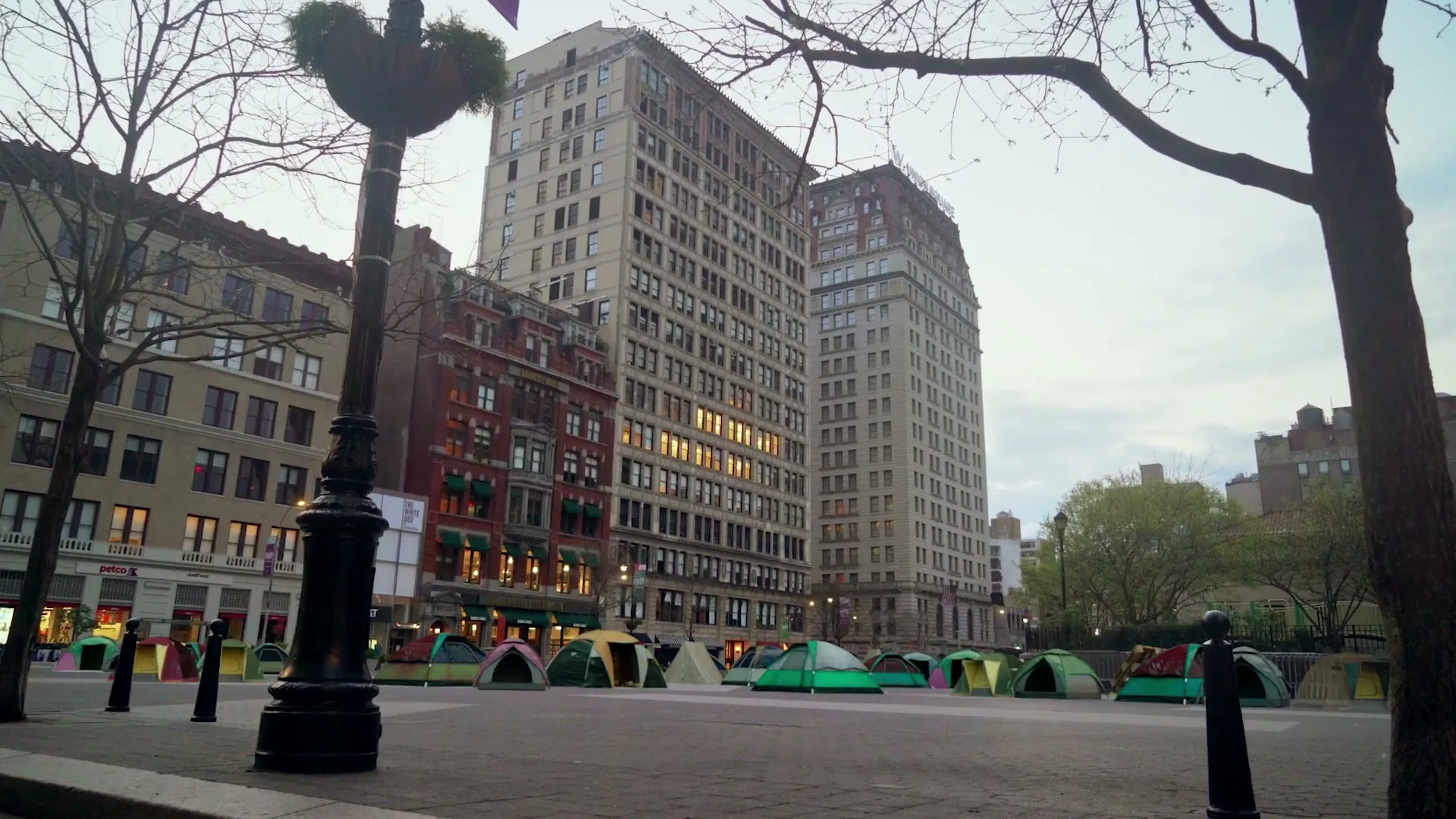 A wide shot of a city square with tall buildings in the background and numerous colorful tents set up on the paved ground, under a cloudy sky.