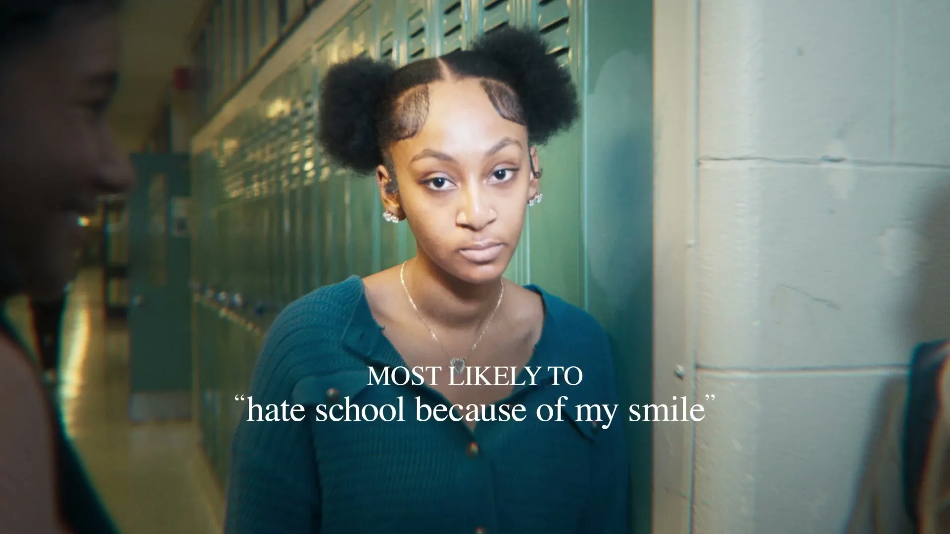 A young Black woman with her hair in two buns stands in a school hallway, looking directly at the camera with a neutral expression.