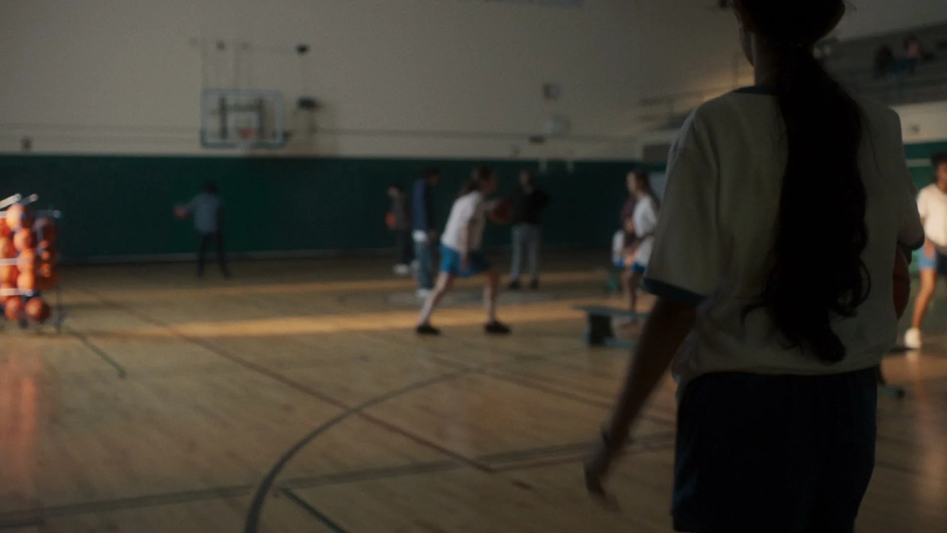 A young female basketball player in a white and blue uniform sits on a bench in a school gym, looking directly at the camera with a neutral expression.