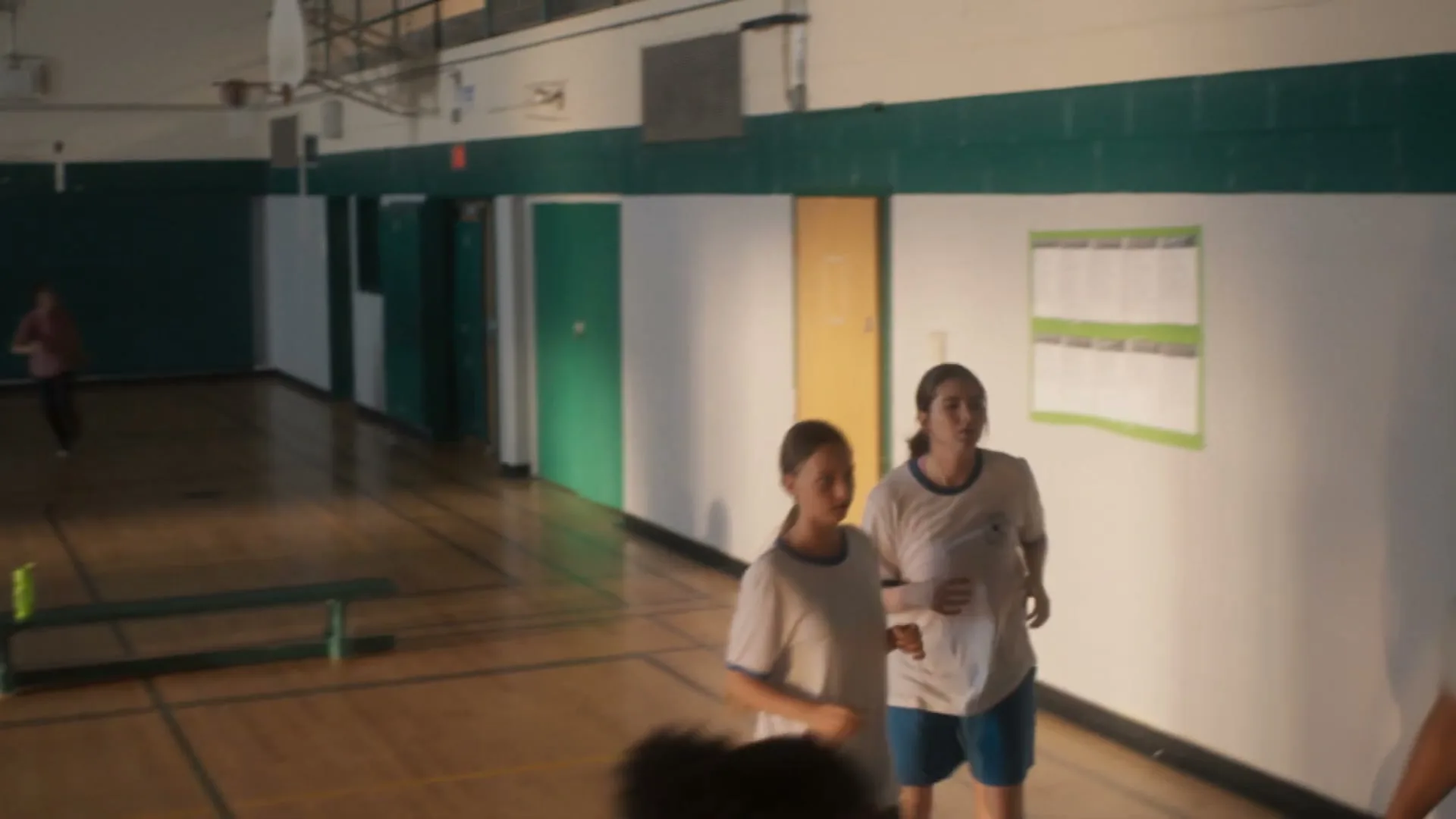 A young woman with dark hair sits on bleachers in a school gym, looking at her phone, then looks up at the camera.