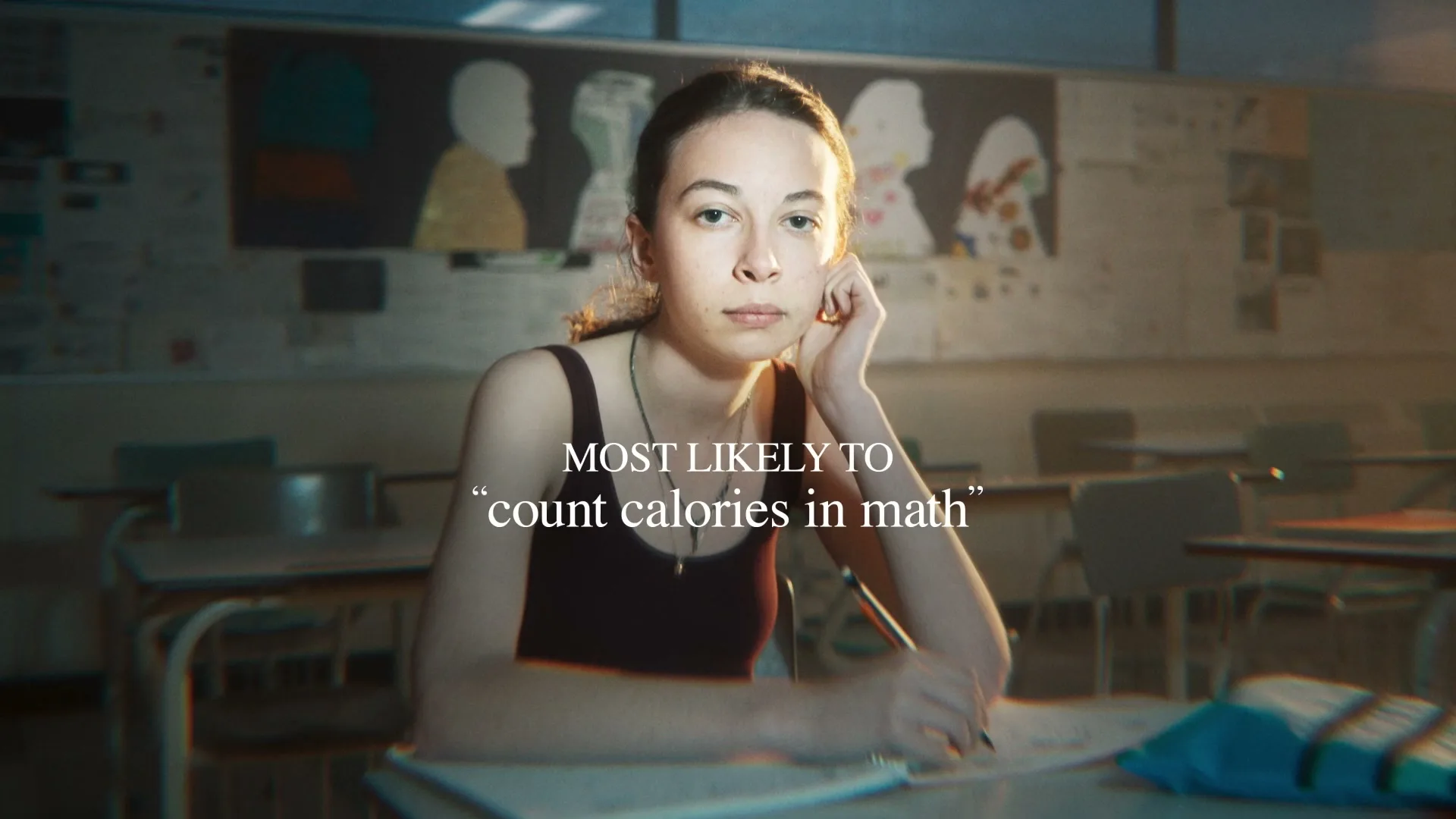 A young female student with dark hair sits at a desk in a classroom, looking directly at the camera with a serious expression.