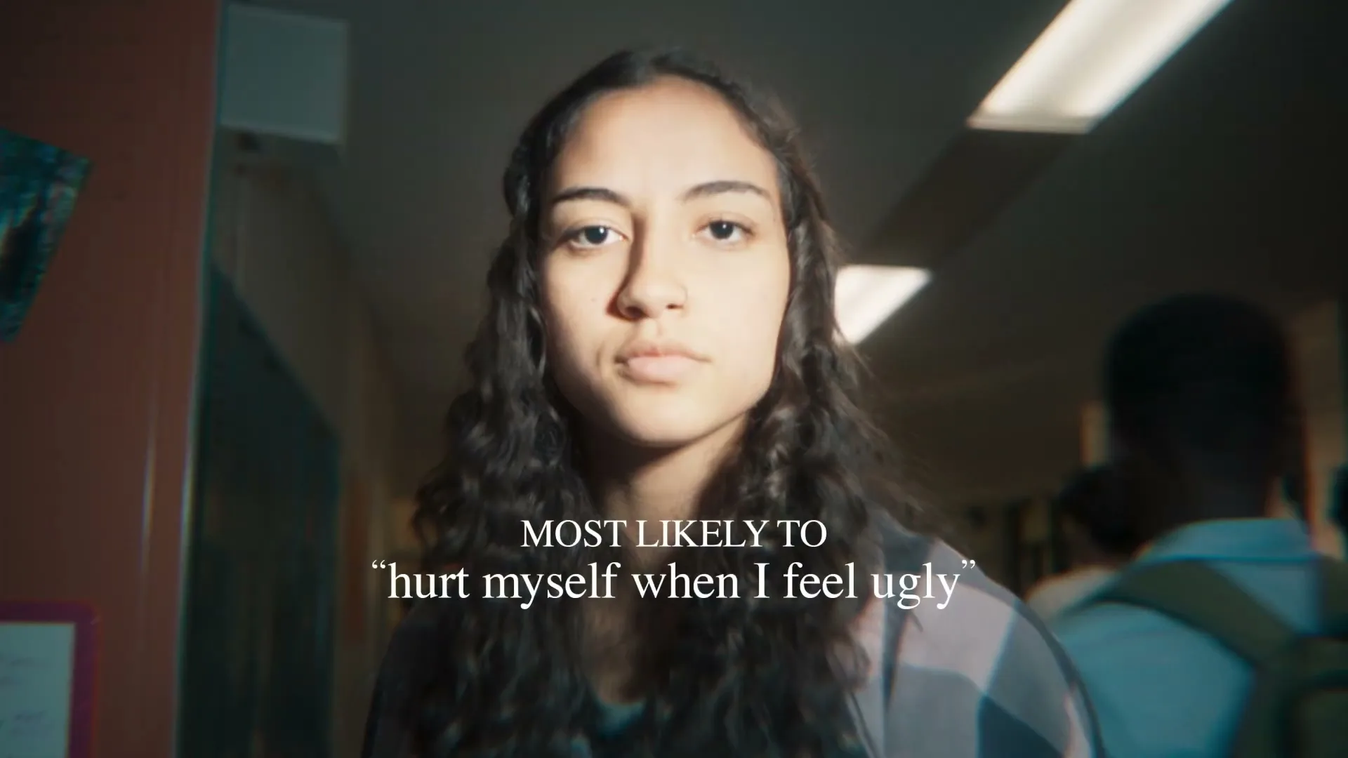 A young female with long curly hair looks directly at the camera with a serious expression in a school hallway, with text overlayed.