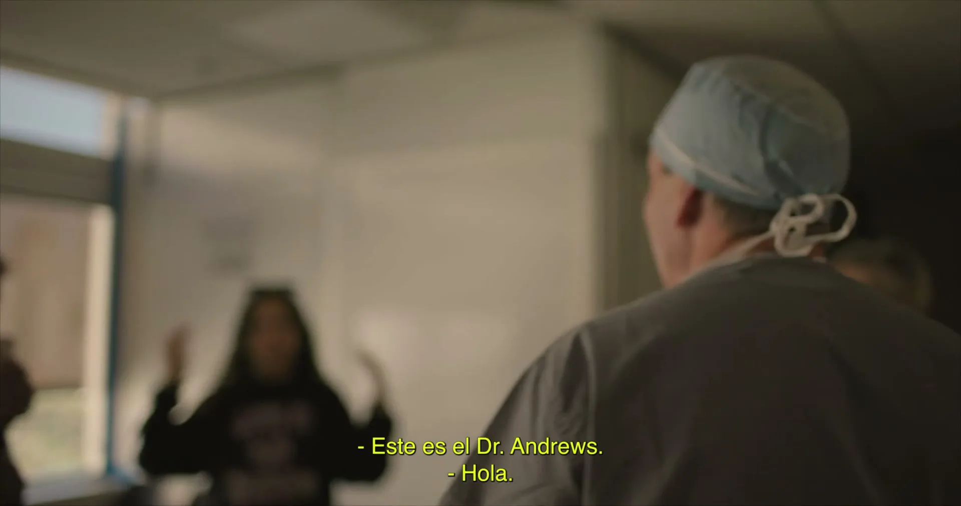 A doctor in scrubs and a surgical cap turns to greet a family, including a baby, in a hospital waiting room.