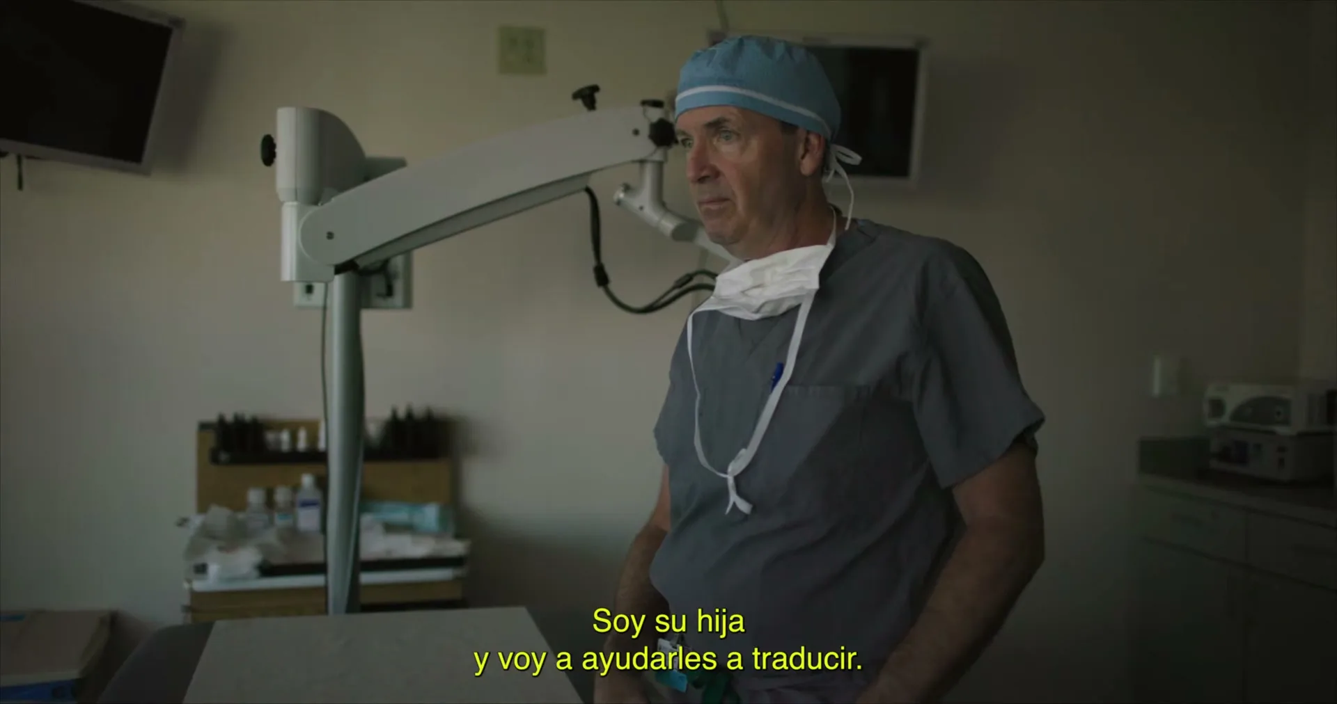 A male surgeon in scrubs and a surgical cap stands in a dimly lit operating room, looking off-camera with a neutral expression.
