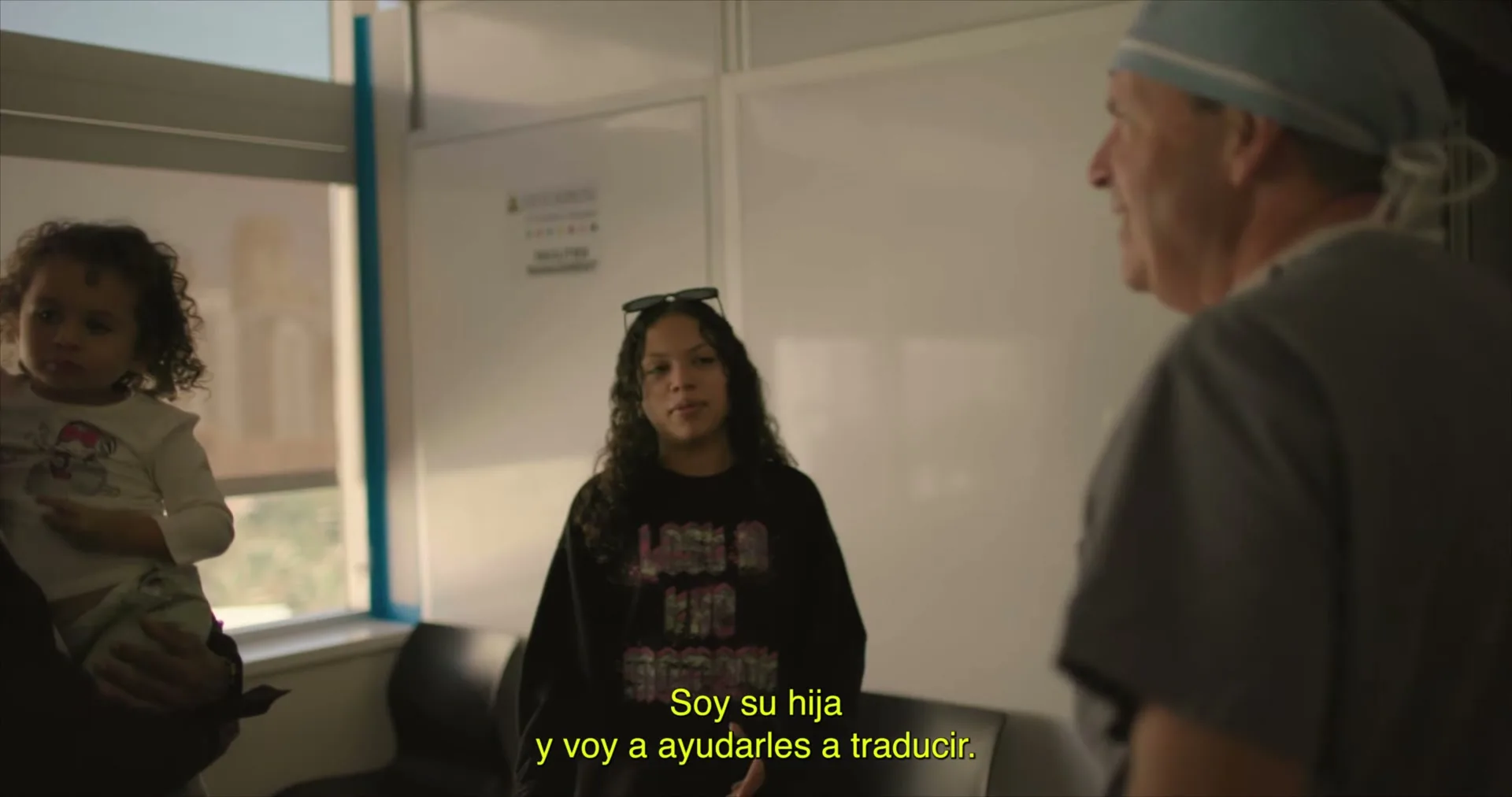 A young girl with curly hair is held by an adult, while a teen girl in a black sweatshirt speaks to a male doctor in scrubs in a hospital room.