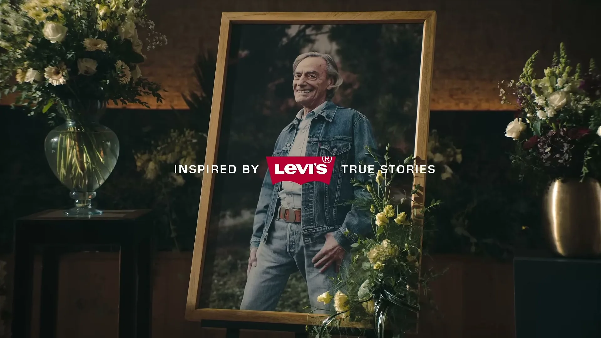 A framed portrait of a smiling older man in a denim jacket and jeans stands surrounded by floral arrangements in a dimly lit interior.