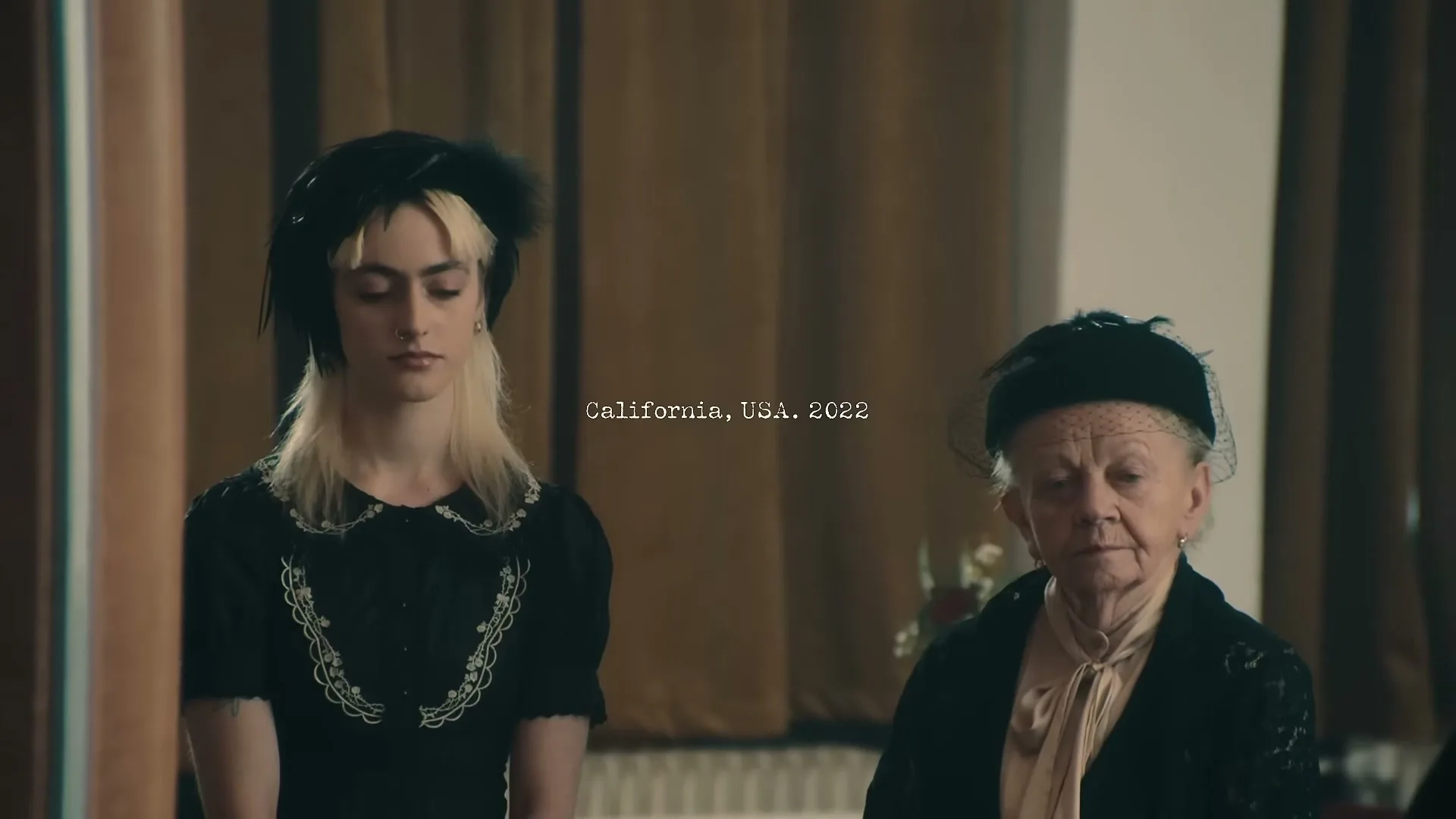A young woman with blonde hair and an older woman in black dresses stand side-by-side in a room with brown curtains, looking forward.