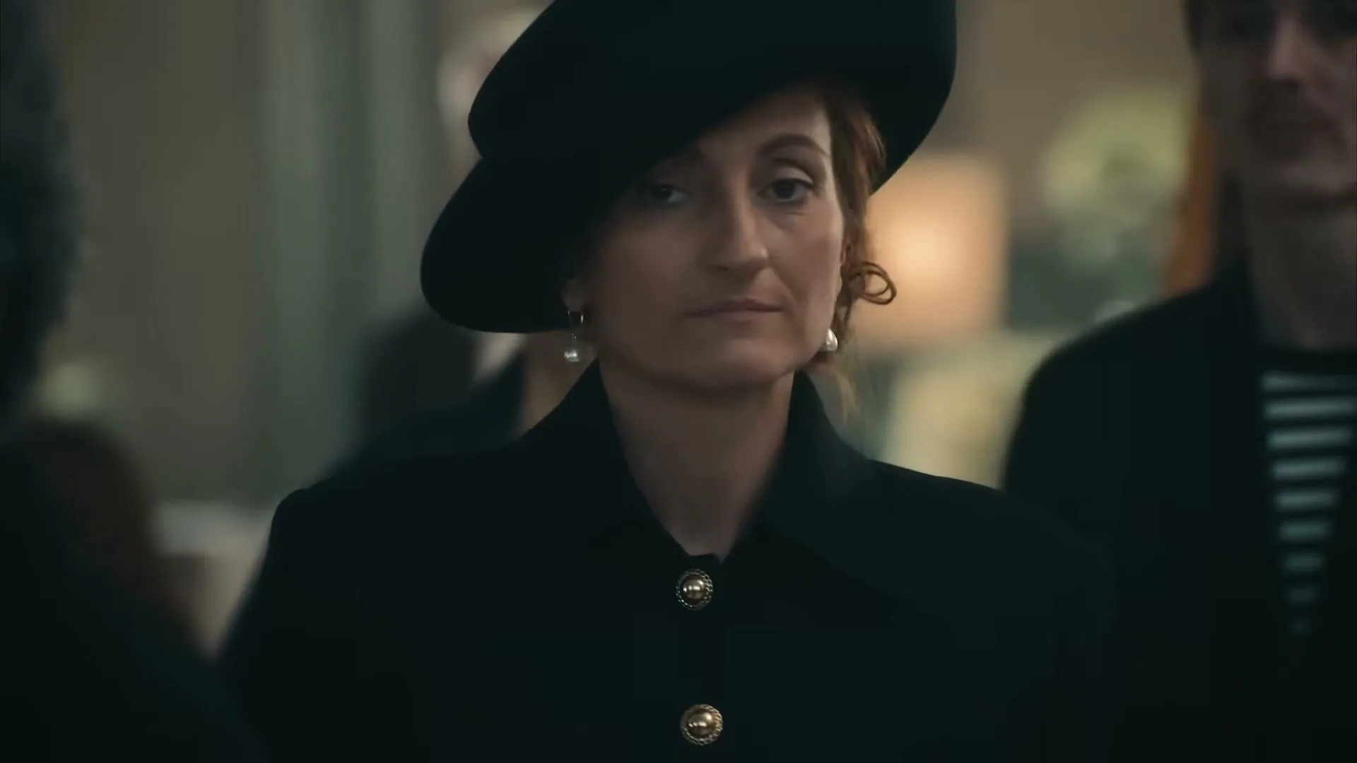 A woman in a black hat and coat with pearl earrings turns her head to face the camera in a dimly lit interior.
