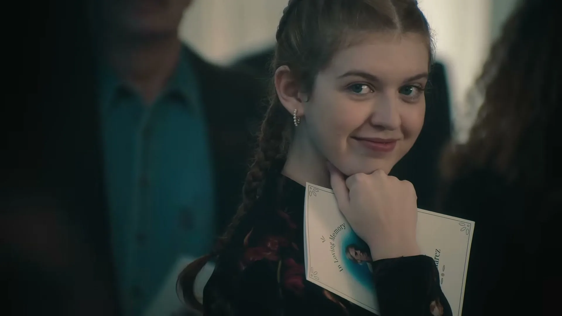 A young woman with braided hair, wearing a dark floral top, holds a memorial program and smiles subtly at a funeral service.