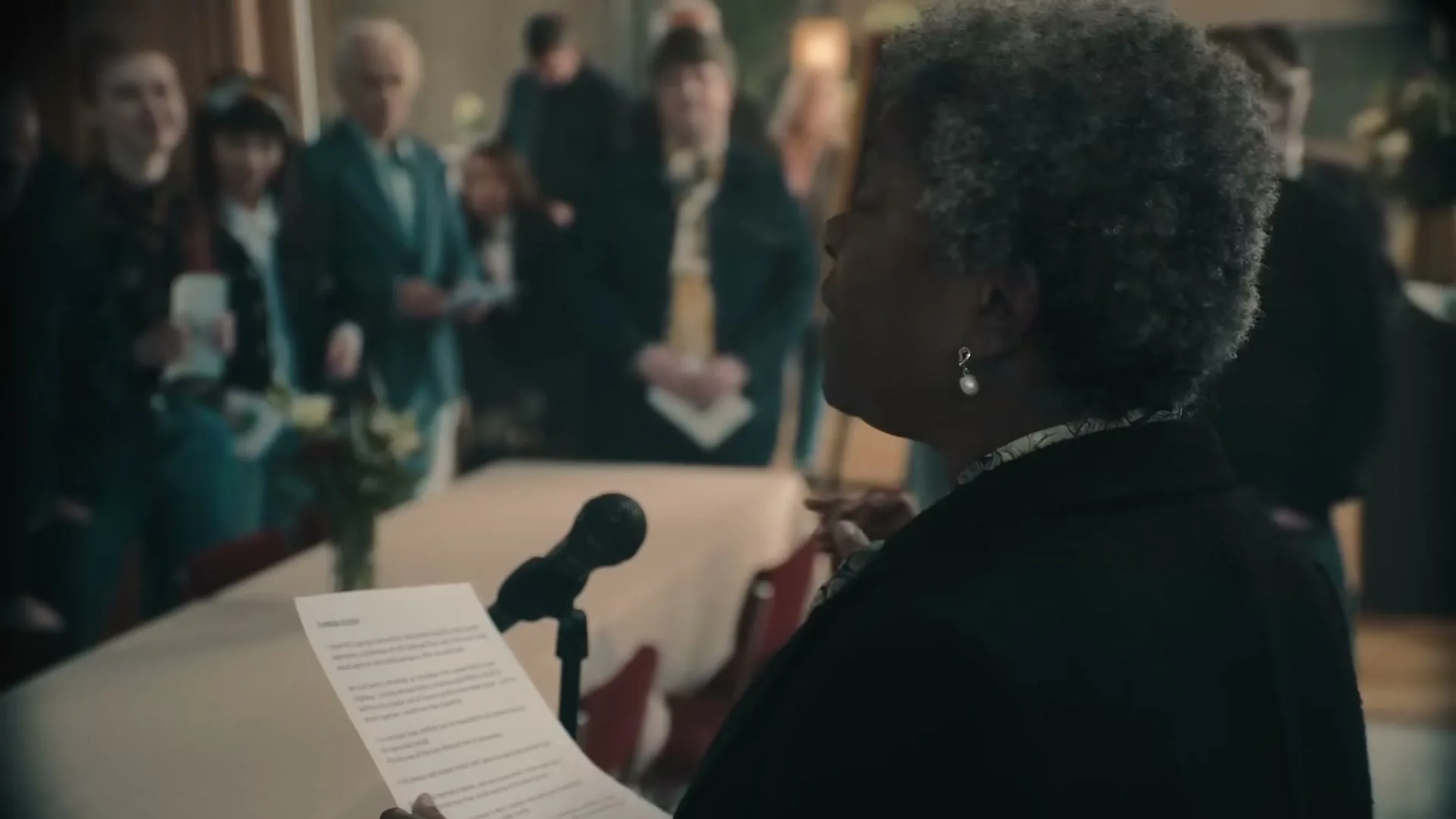 An older Black woman with grey hair and pearl earrings stands at a podium, reading from a paper to a small group of people in a room.