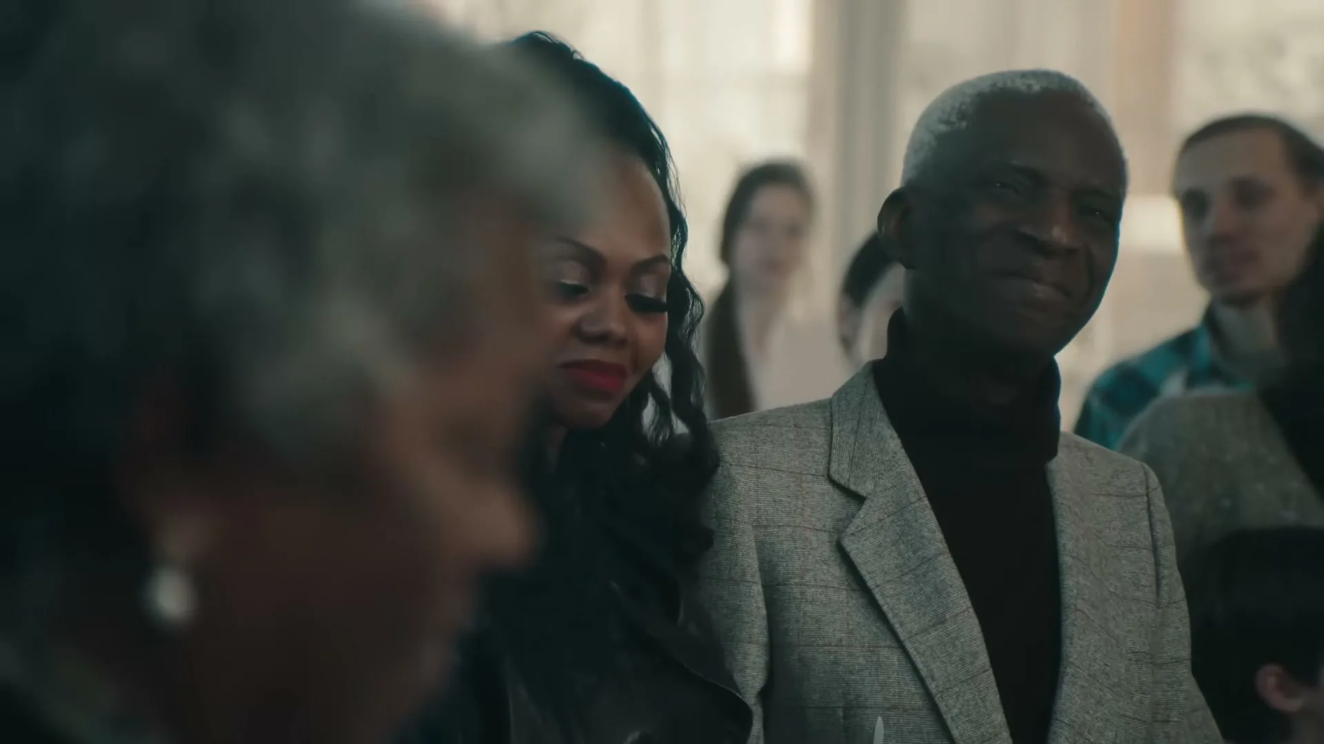 A diverse group of people, including an older Black man in a tweed jacket and a younger Black woman with long dark hair, are gathered indoors, looking forward.