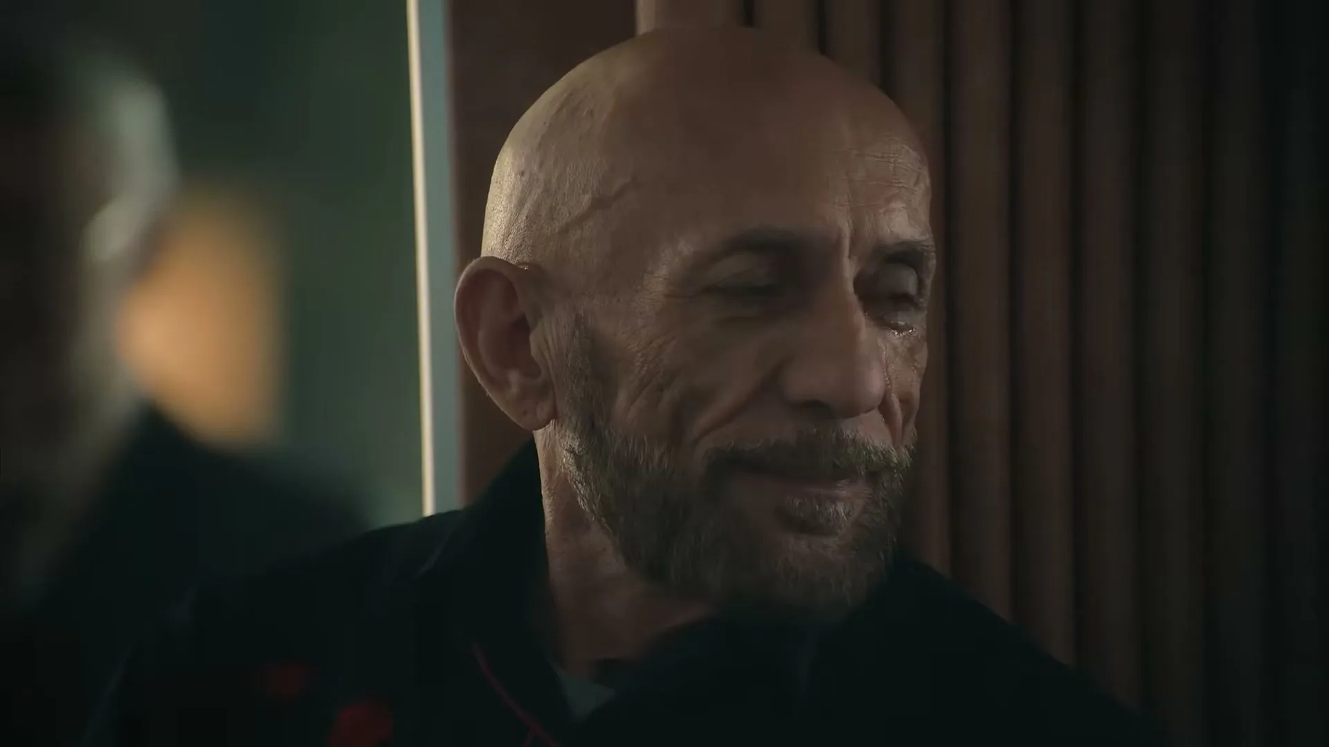 A bald, bearded man in his 50s or 60s, wearing a dark jacket, looks off-camera with a slight smile in an interior setting.