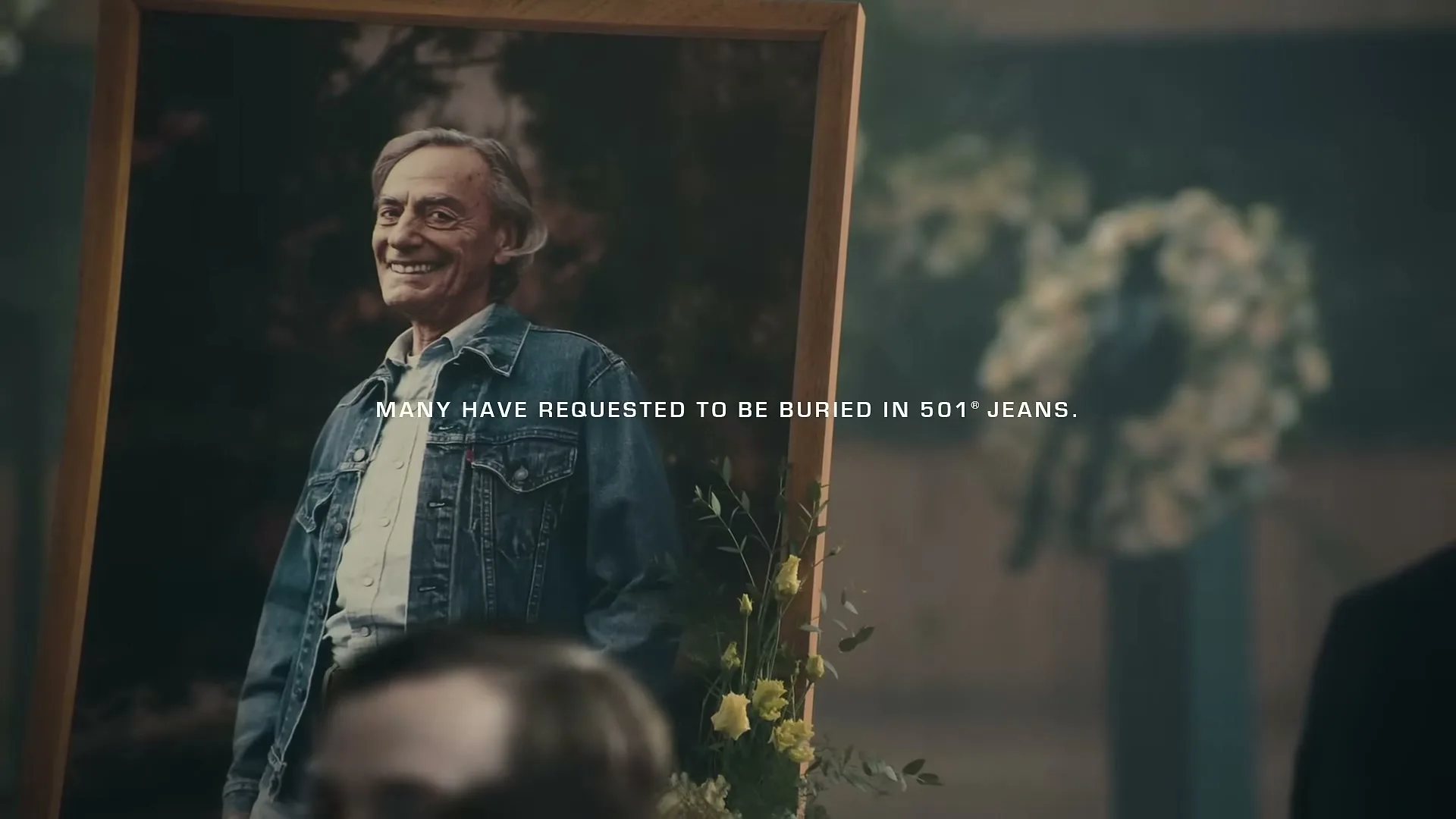 A framed portrait of a smiling older man in a denim jacket is displayed at a funeral, with a young man in a suit visible in the foreground.