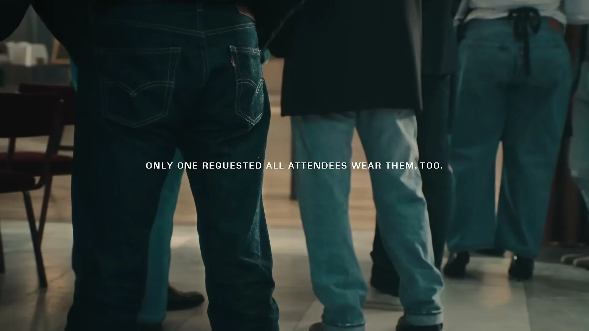 A group of people, mostly men, are seen from behind, standing in an interior space, all wearing jeans.