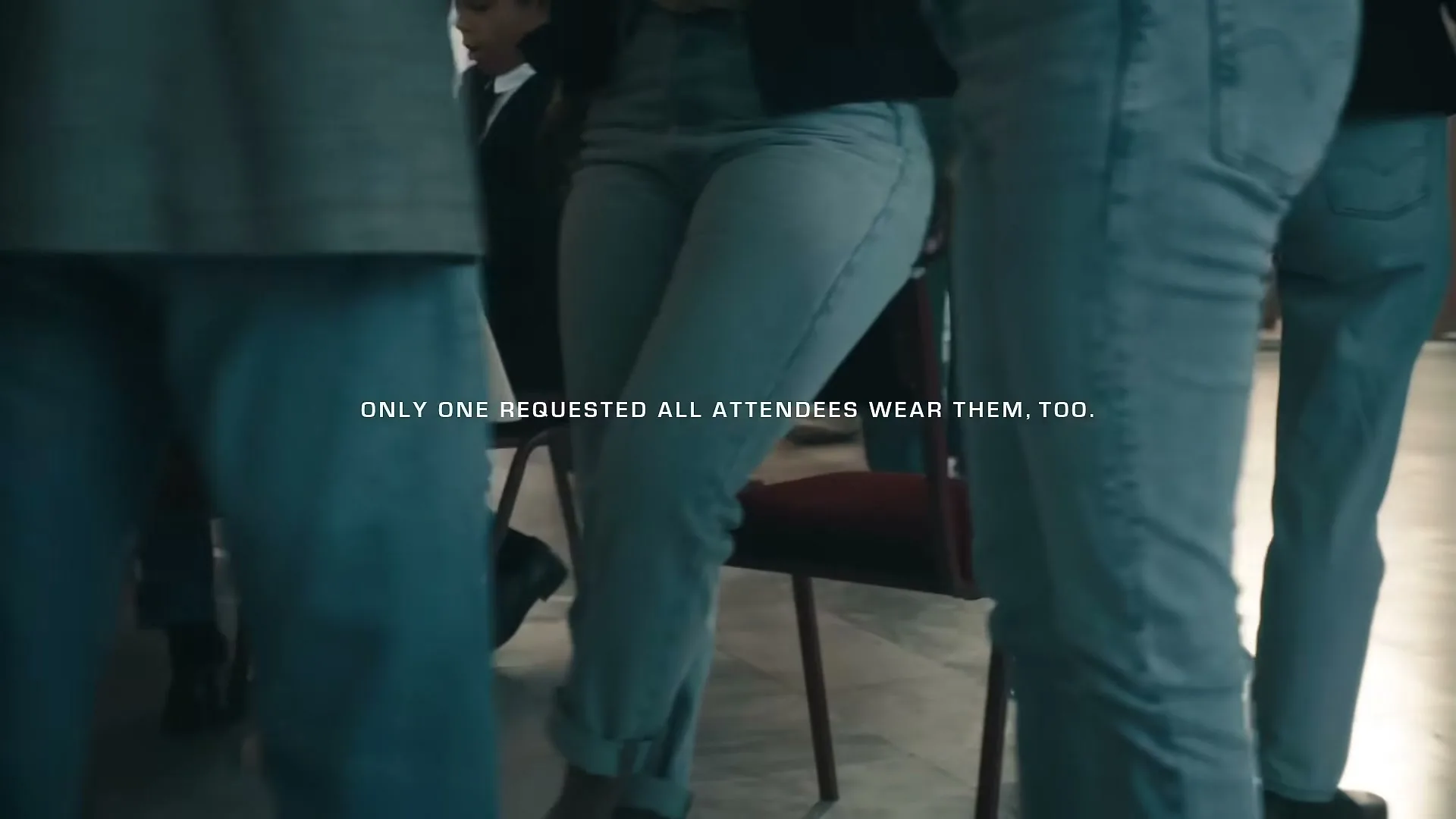 A group of people wearing blue jeans are standing and moving around in an interior setting, with chairs visible in the background.