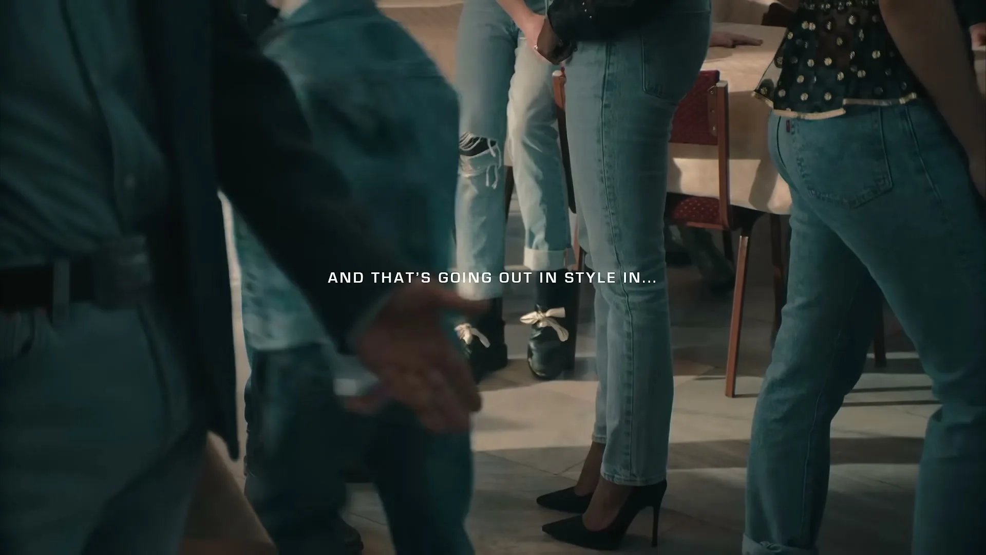 A group of people wearing various styles of blue jeans and different shoes are standing around a table in an interior setting.