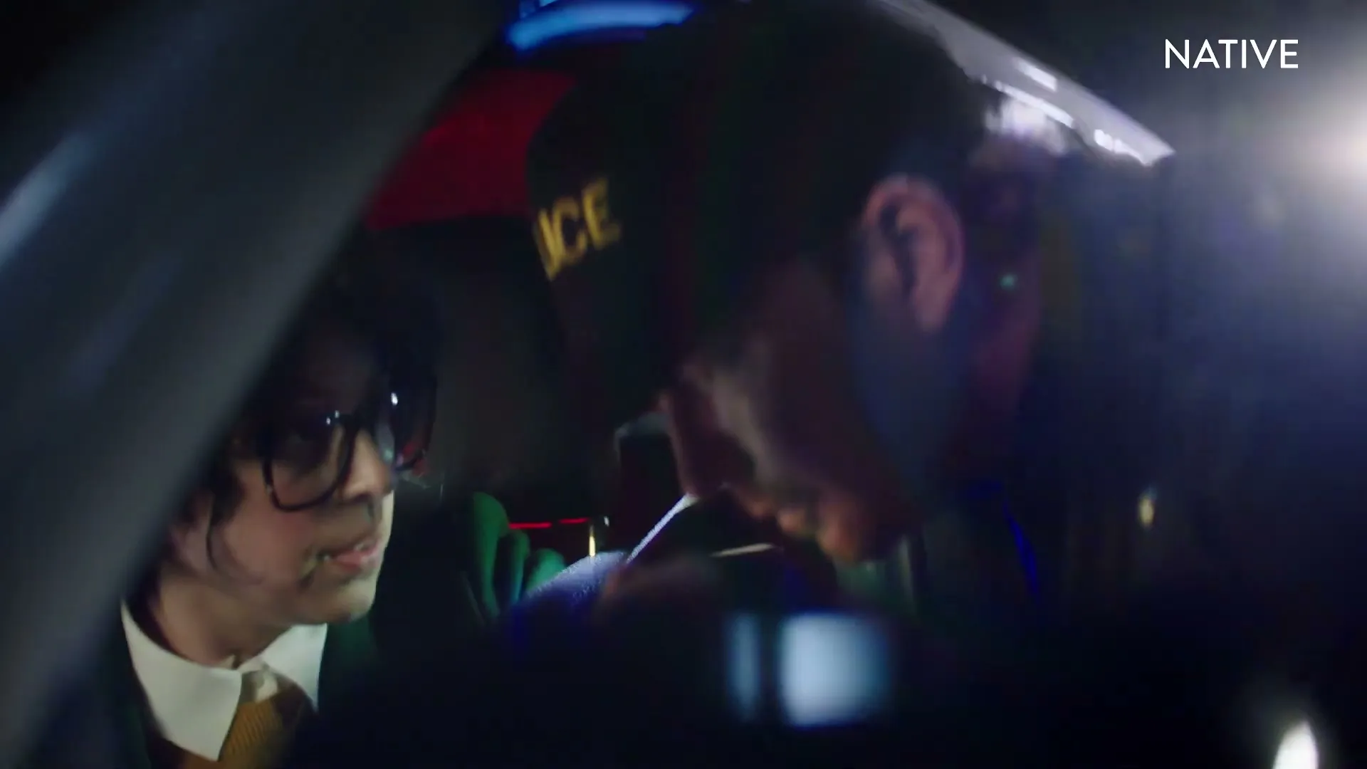 A female driver with glasses looks up at a male police officer leaning into her car during a nighttime traffic stop.