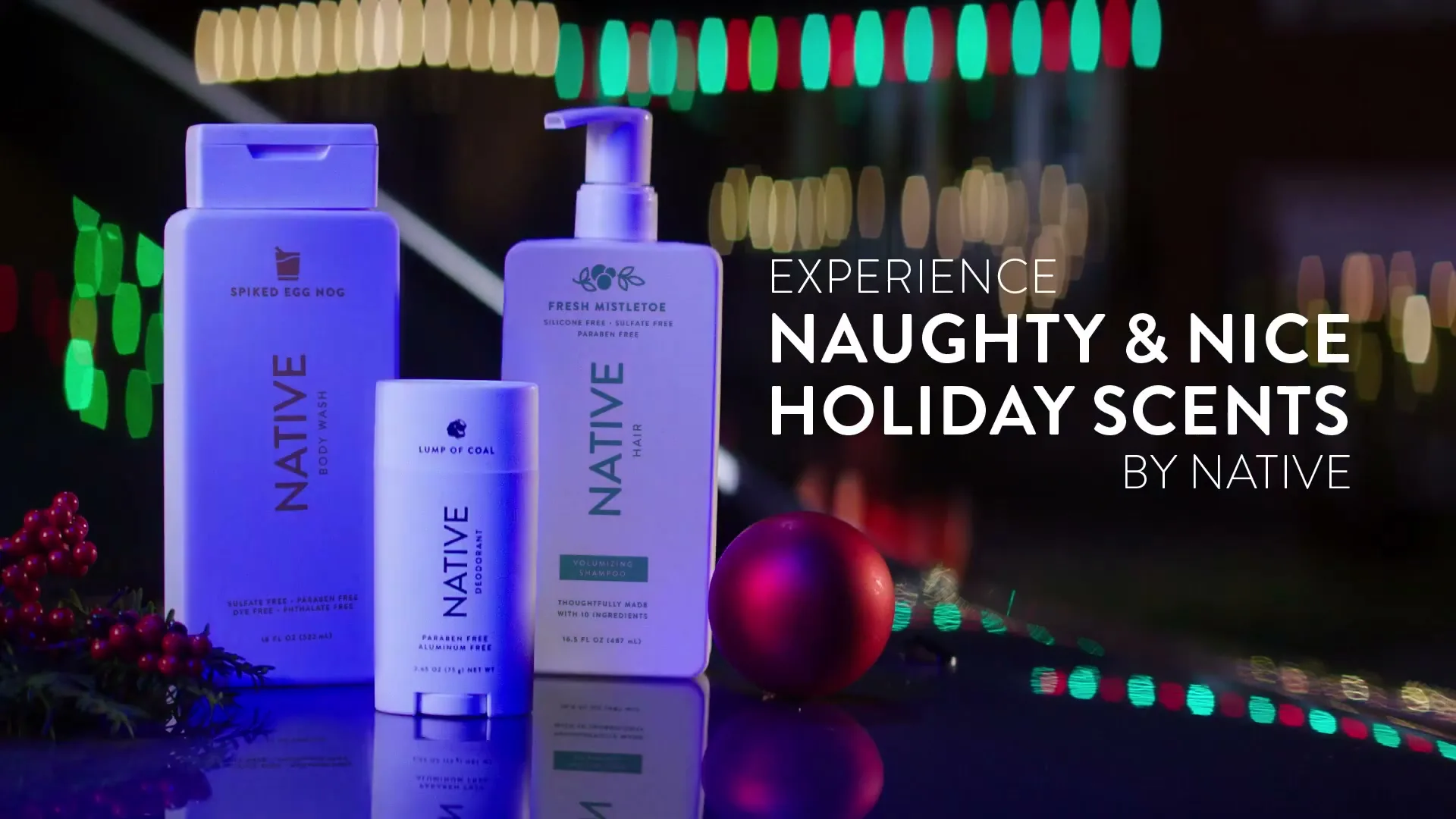 Three Native brand holiday-themed personal care products are displayed on a reflective surface with blurred festive lights in the background.