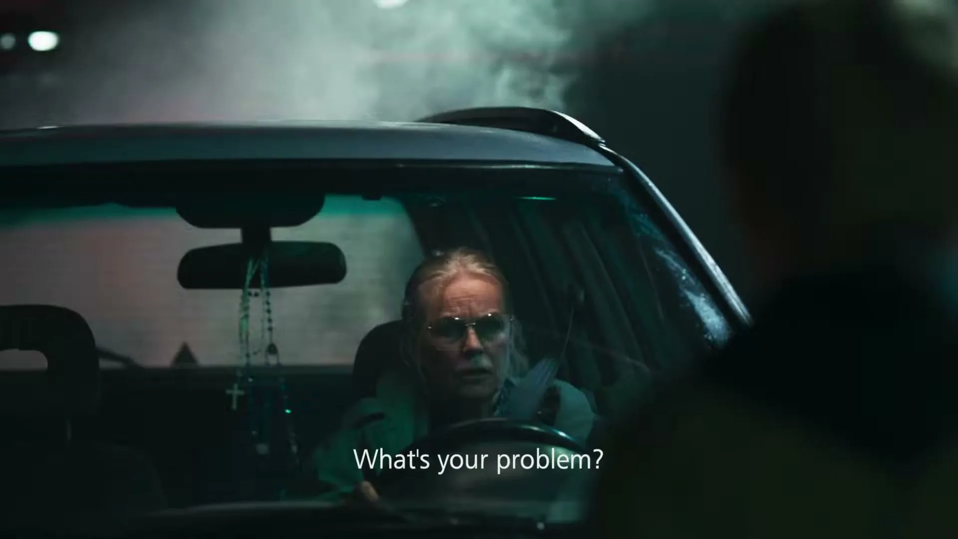 An older woman with glasses and a teal jacket yells from inside her car at an unseen person, her face showing anger and frustration.