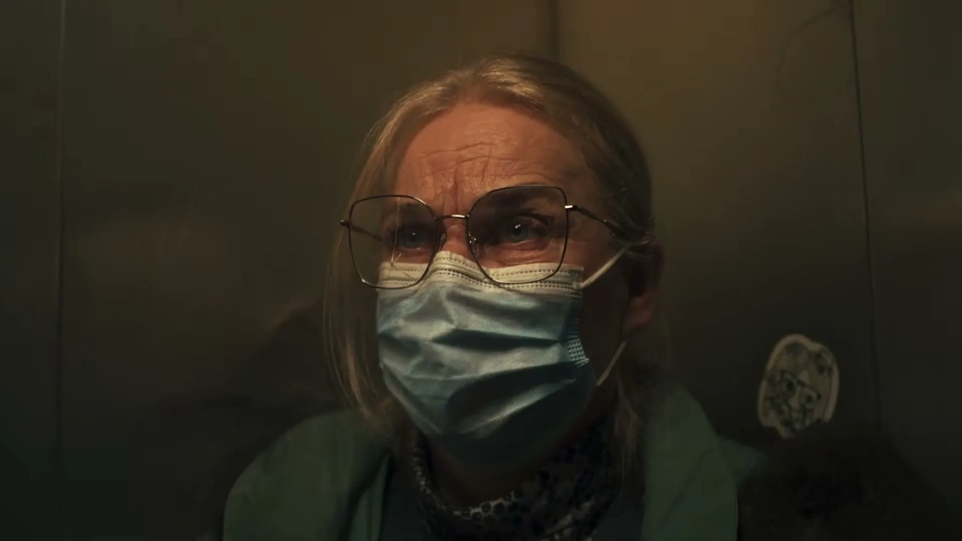 A senior woman with blonde hair, glasses, and a blue face mask looks up with a worried expression in a dark, metallic elevator.