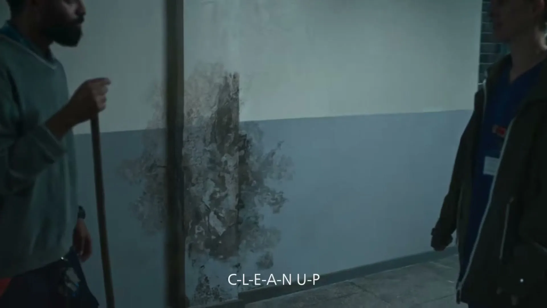 A man and a woman stand in a dimly lit hallway, looking at a wall with peeling paint and water damage, as the woman points at the wall.