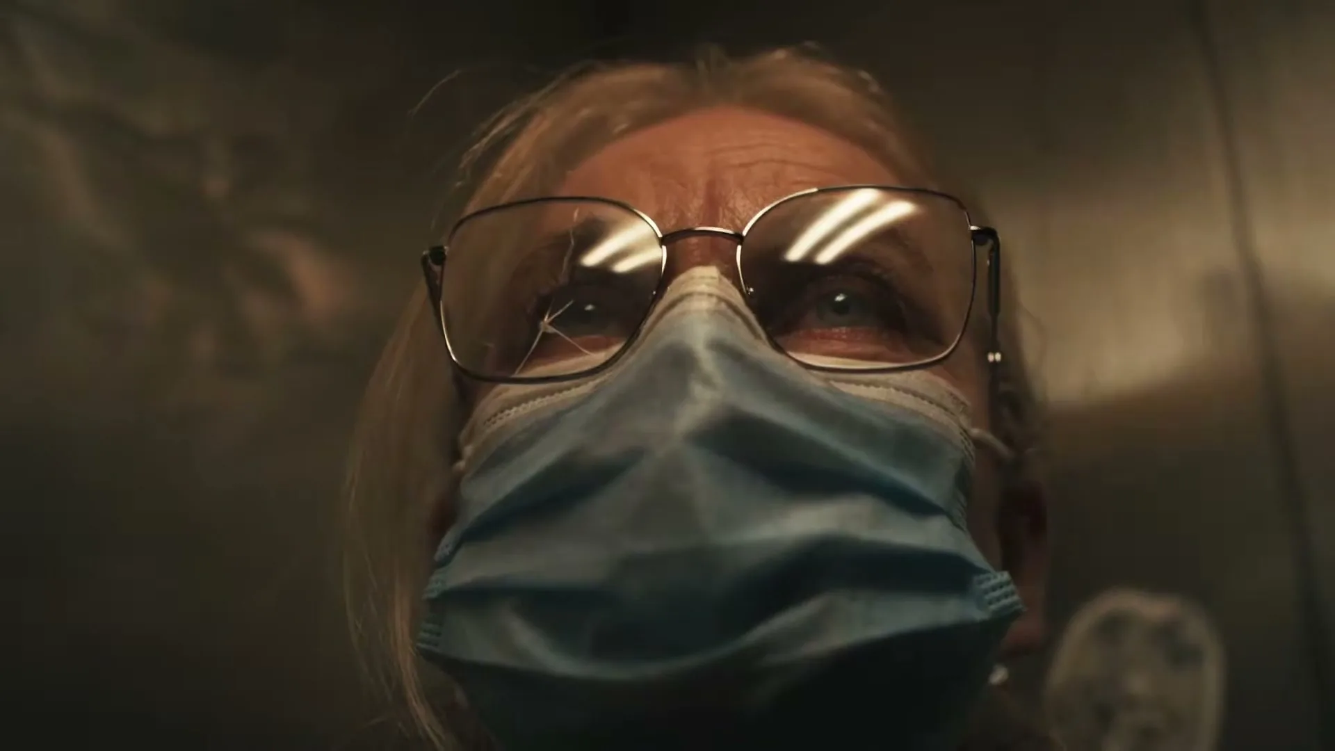 A close-up of an older woman with blonde hair, glasses, and a blue medical mask, looking up with a concerned expression in an elevator.