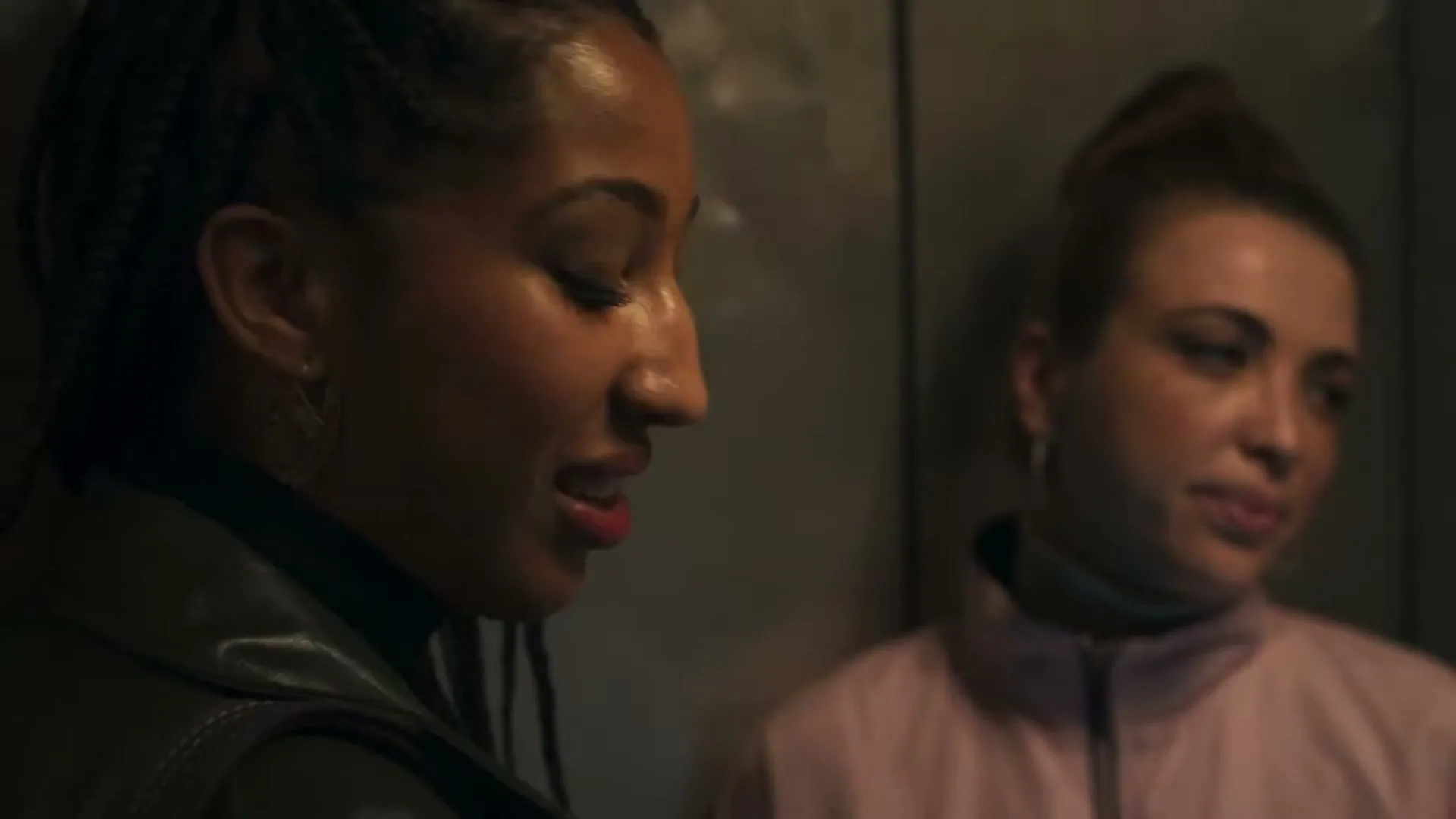 Two young women, one with braids and a green jacket, the other in a pink top, stand close together in an elevator, looking downcast.
