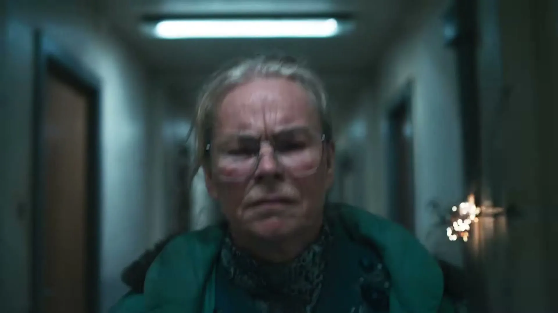 An older woman with glasses and a green jacket looks distressed while walking down a dimly lit, narrow hallway.