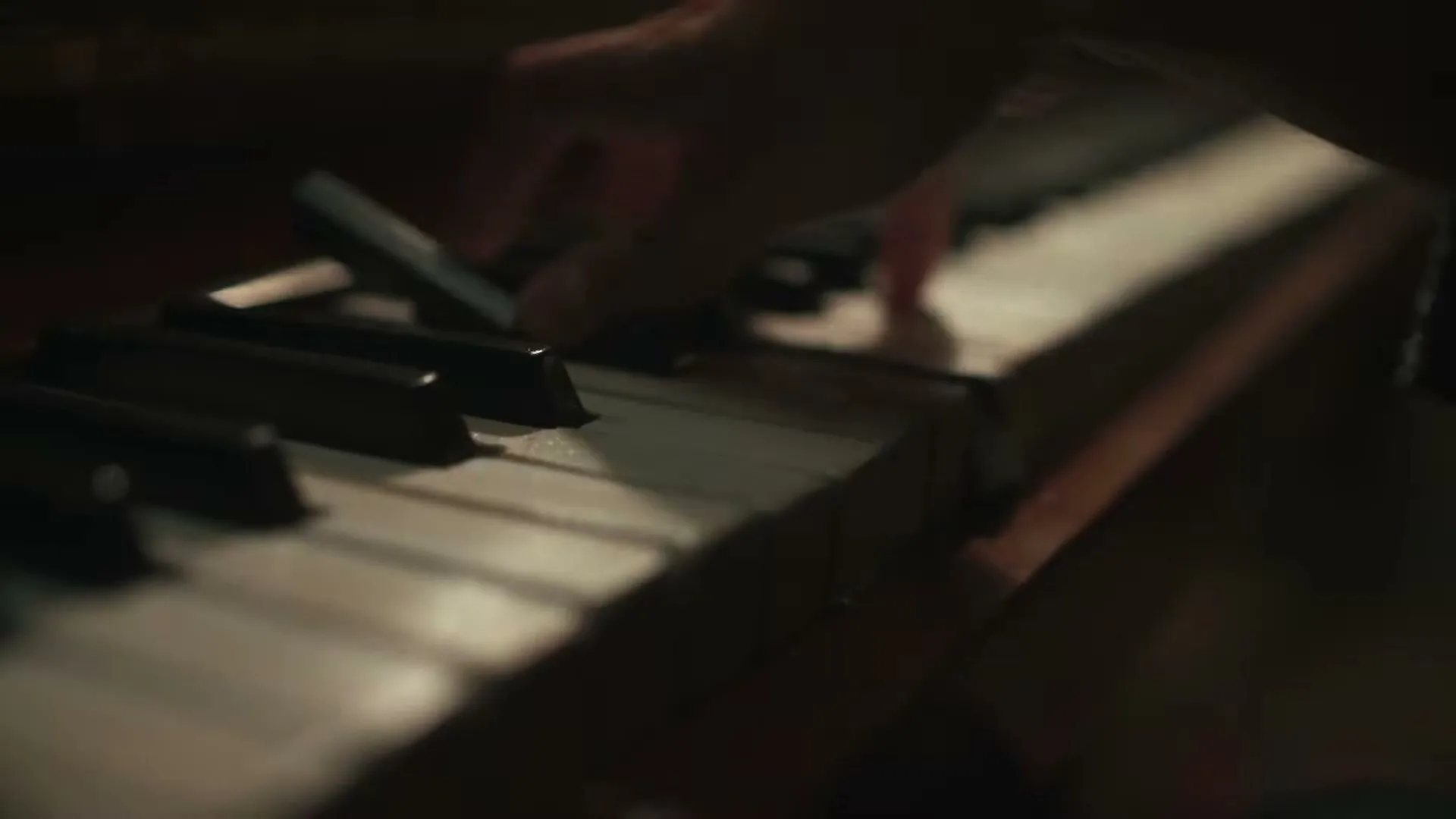 A person's hand plays a melody on the keys of an old piano, dimly lit in a dark room.