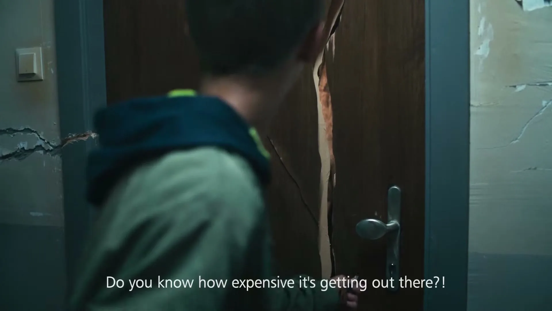 A young man in a hoodie looks at a wooden door with a large crack in it, in a dimly lit hallway.