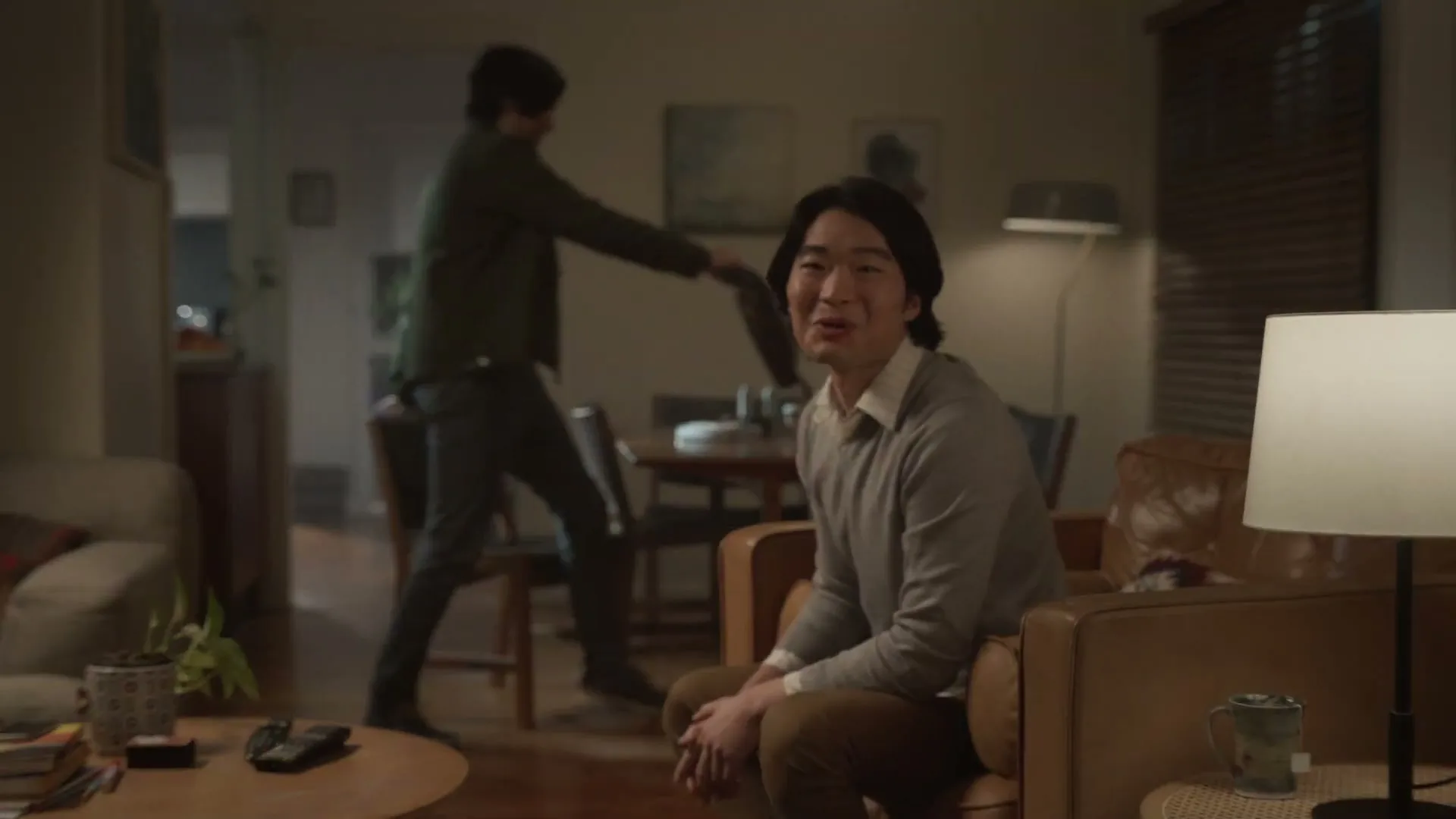 Two men are in a dimly lit living room, one sitting on a couch and the other walking towards a dining table, both smiling.