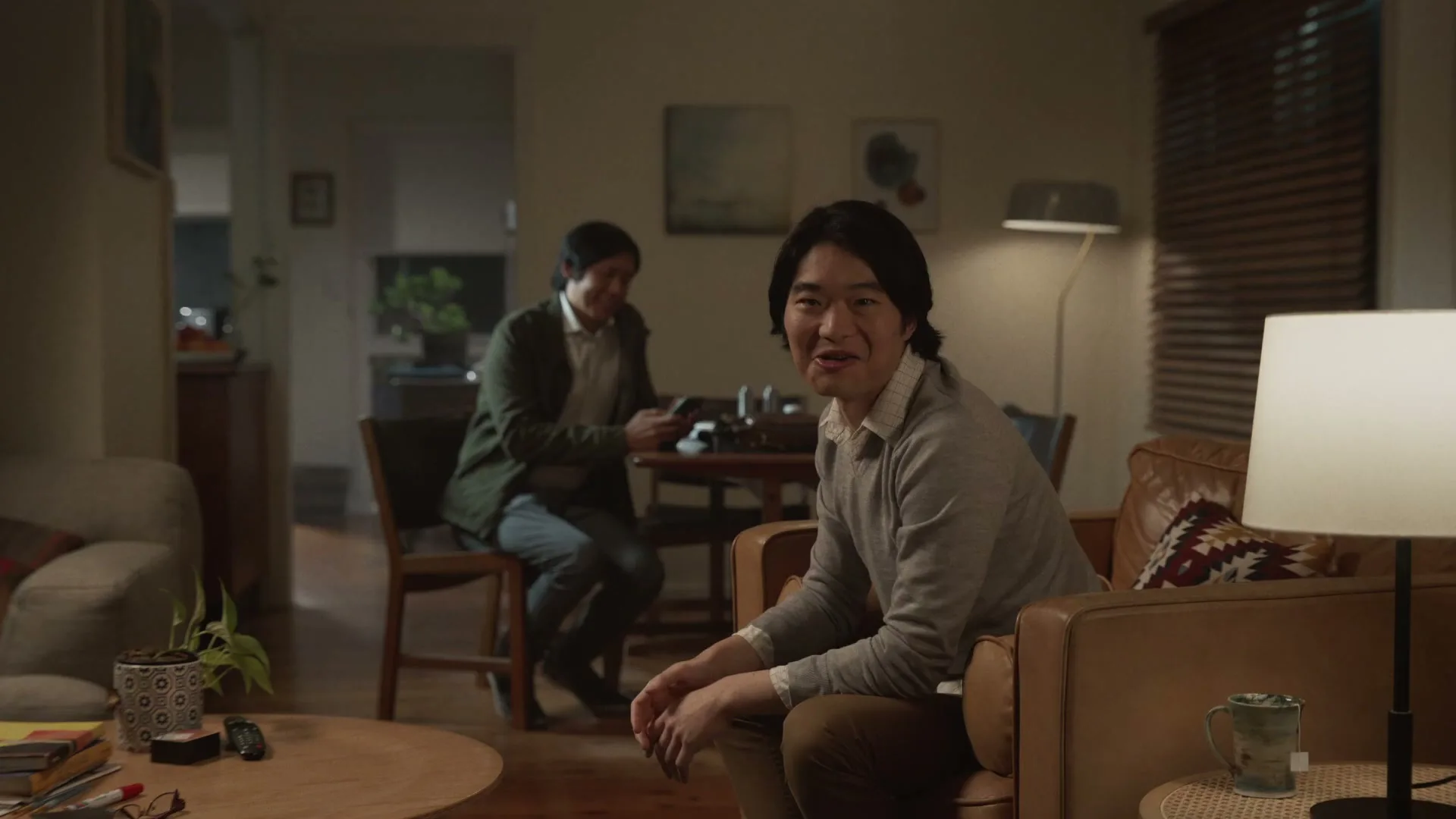 Two men are sitting in a living room, one on a couch and the other at a dining table, looking at each other and smiling.