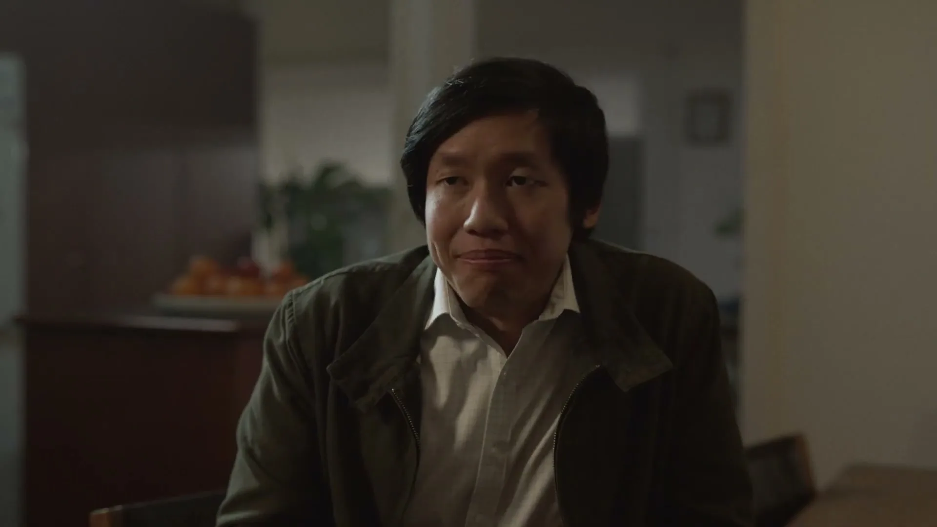A man with dark hair and a white shirt under a dark jacket looks down and then up, appearing to be in a home interior.