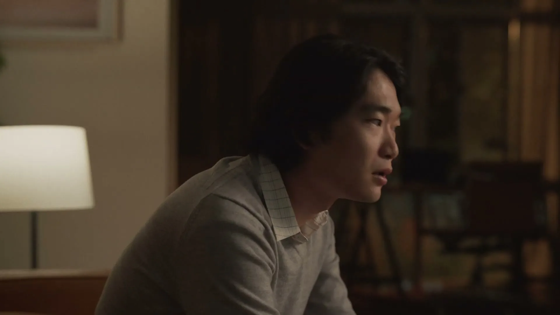 A young Asian man with long dark hair, wearing a grey sweater, looks around with a surprised expression in a dimly lit living room.