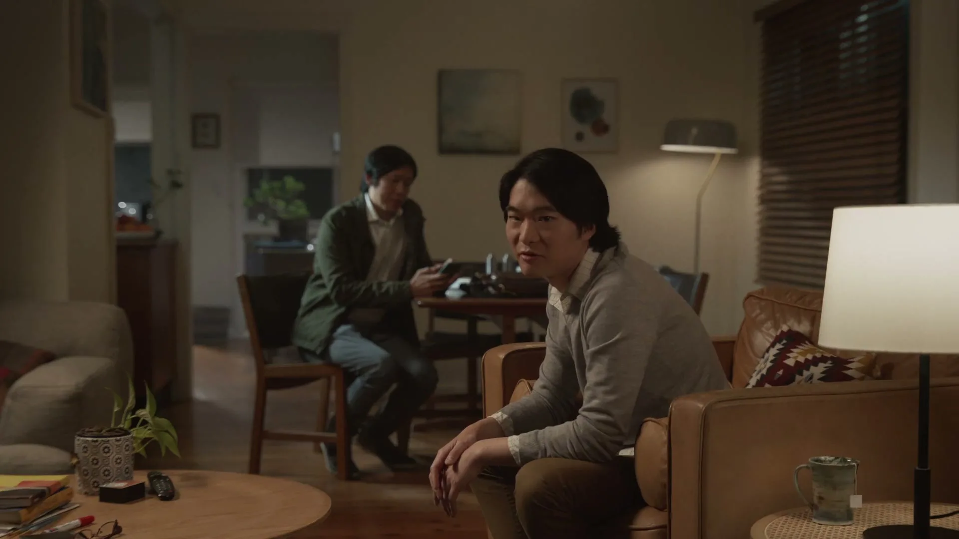 Two Asian men are in a dimly lit living room, one on a couch and the other at a dining table, as the man on the couch looks at the camera.