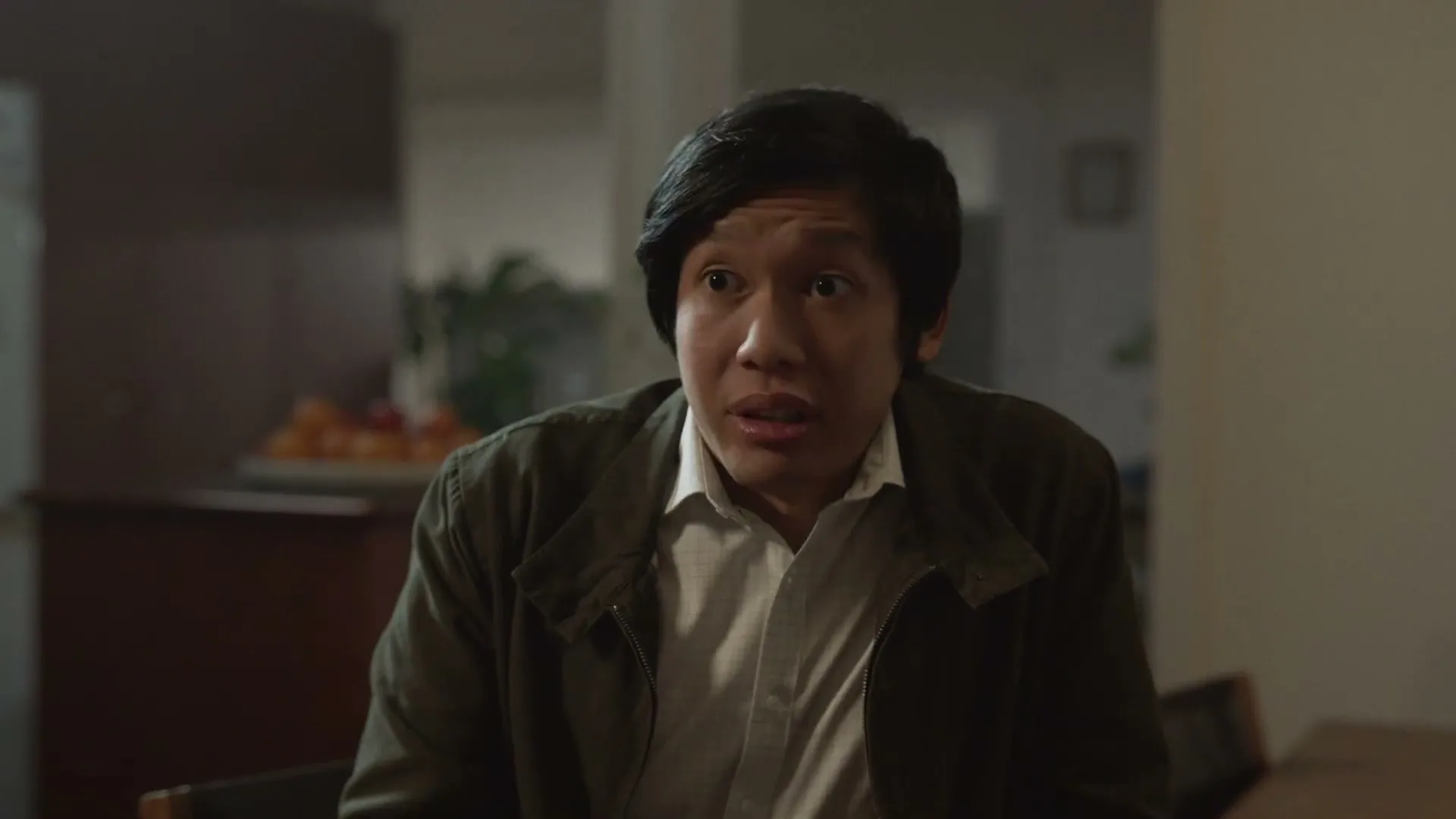 A man with dark hair and a green jacket looks up in surprise while sitting at a table in a dimly lit room.