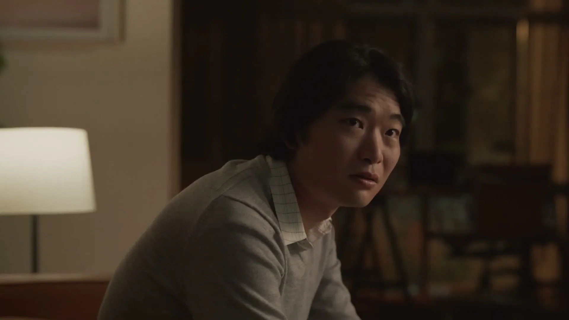 A young Asian man with long dark hair, wearing a grey sweater, looks around with a confused expression in a dimly lit room.
