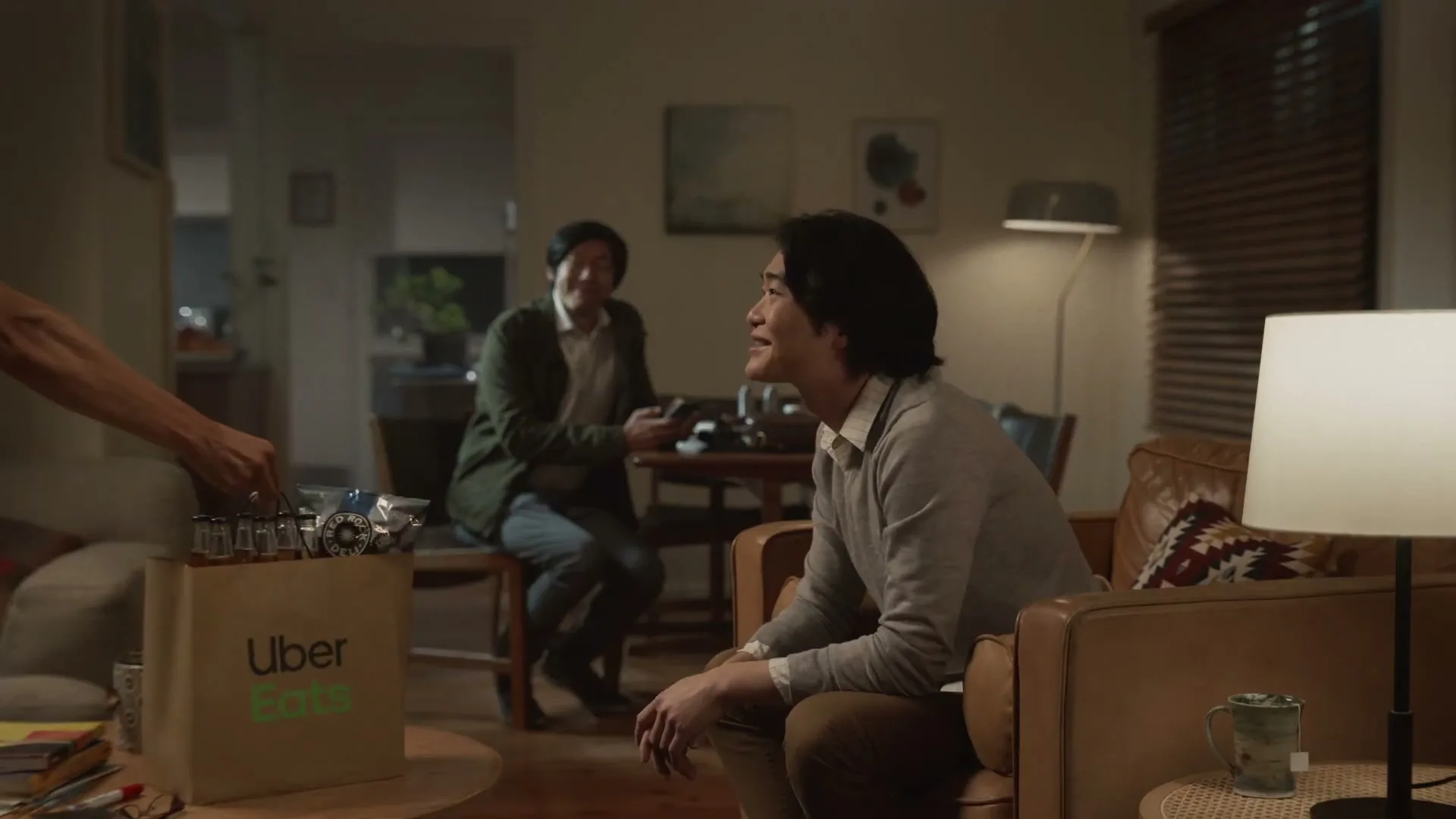 A man sitting on a couch receives an Uber Eats delivery bag from an unseen person, while another man sits at a table in the background.