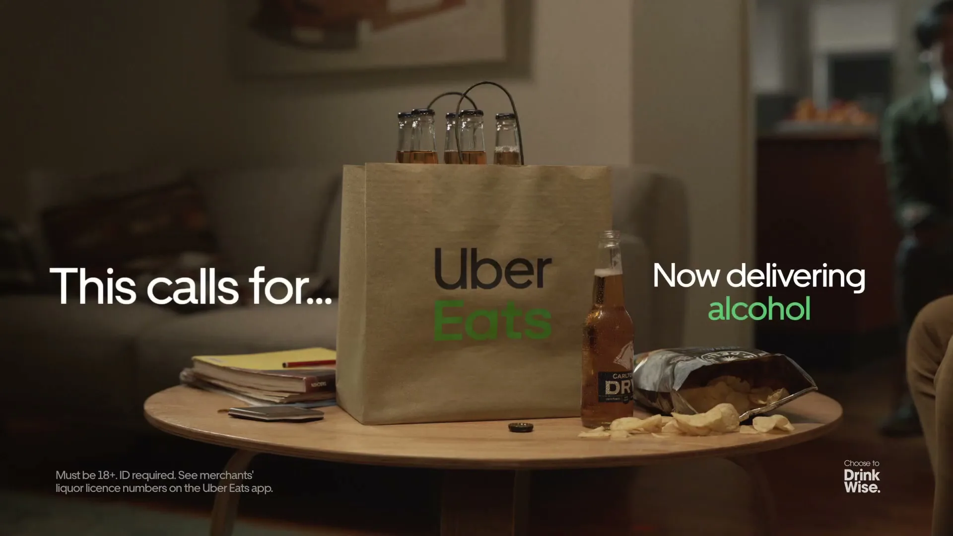 A brown Uber Eats paper bag with four beer bottles and a bag of chips sits on a coffee table in a living room.