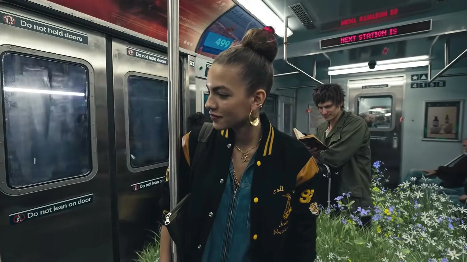 A young woman with a bun and a varsity jacket stands in a subway car, looking to her right, while a man reads a book in the background.