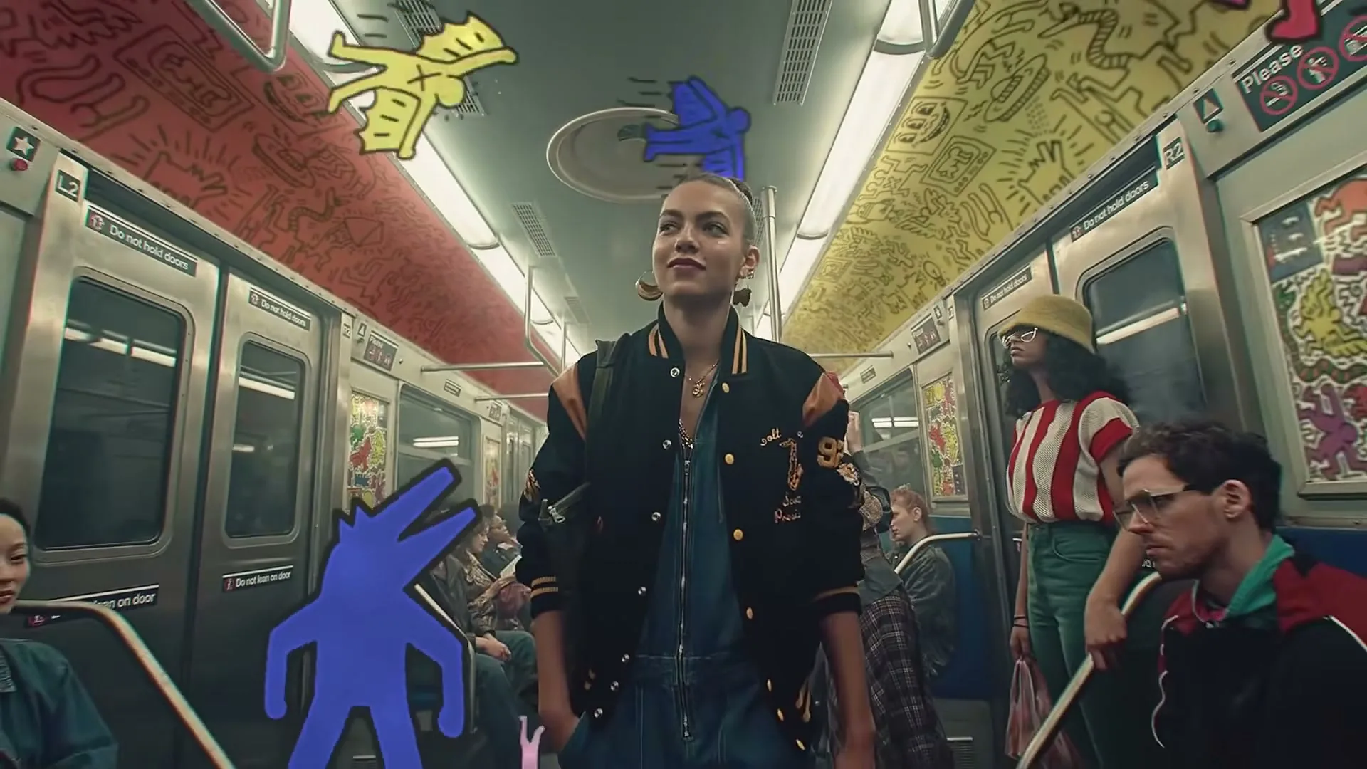 A young woman in a varsity jacket stands in a subway car filled with diverse passengers and animated Keith Haring-style figures.
