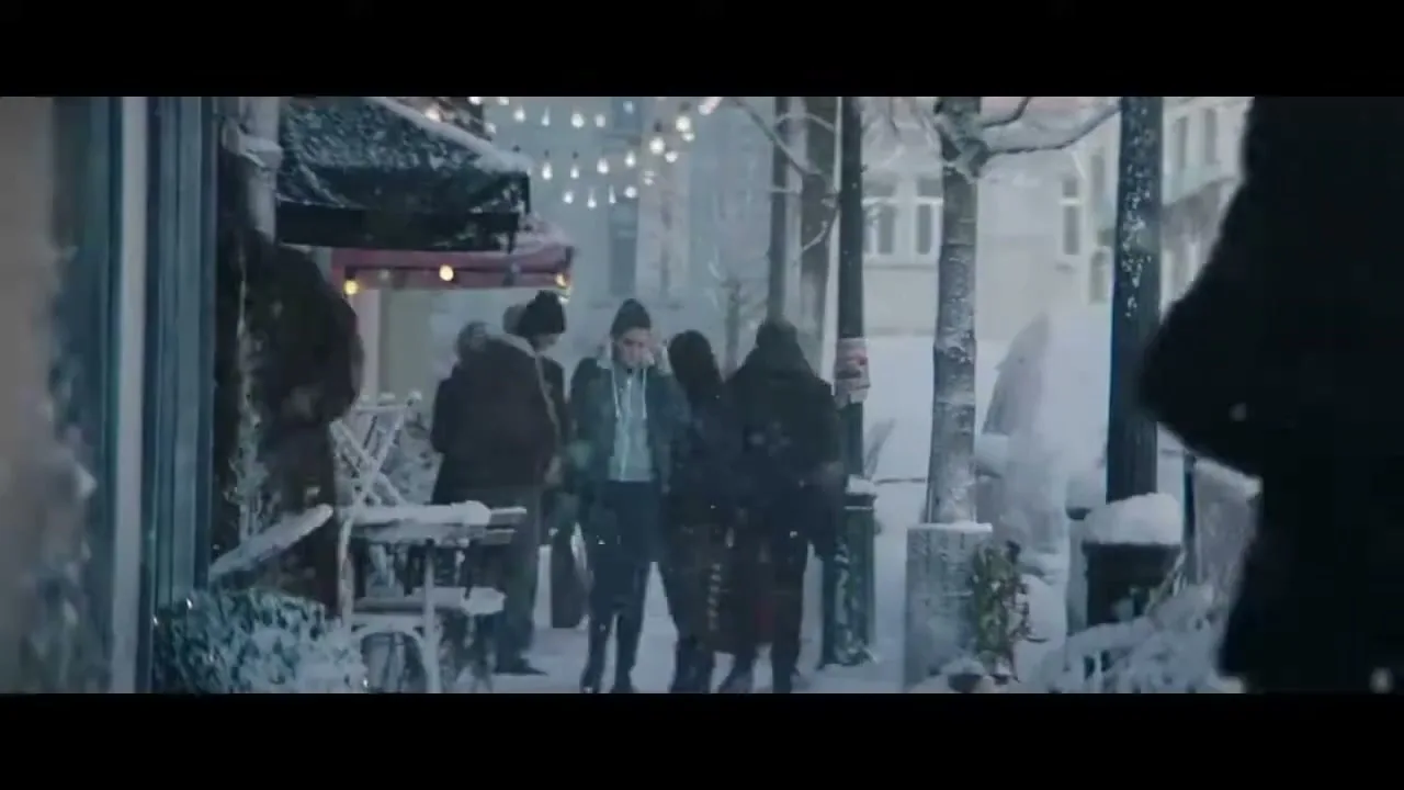 A young woman in a winter coat walks down a snowy city street, surrounded by other pedestrians and storefronts, looking around with a neutral expression.