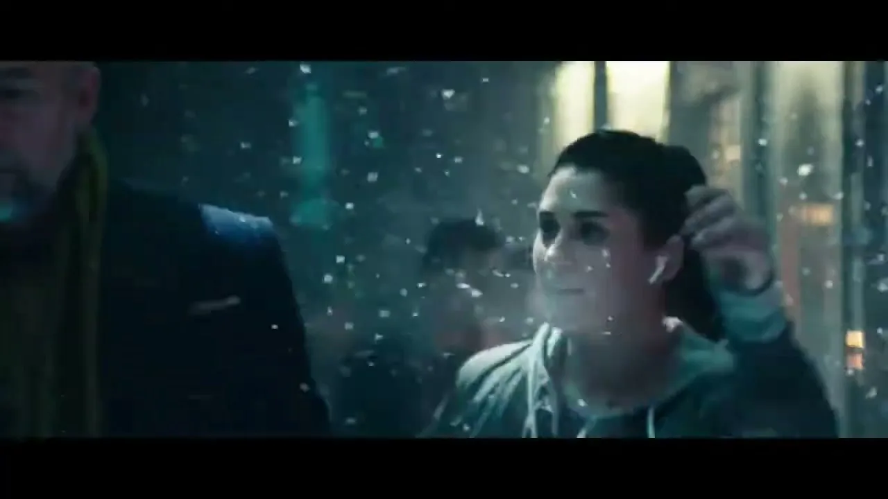 A young woman with dark hair in a ponytail, wearing a light blue hoodie and white AirPods, dances joyfully in a snowy urban street at night.