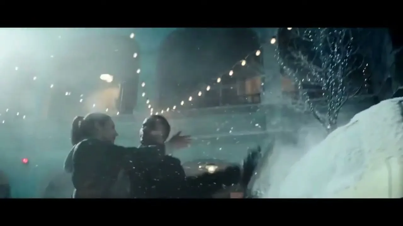 A man and woman dance playfully in a snowy town square at night, illuminated by string lights and falling snow.