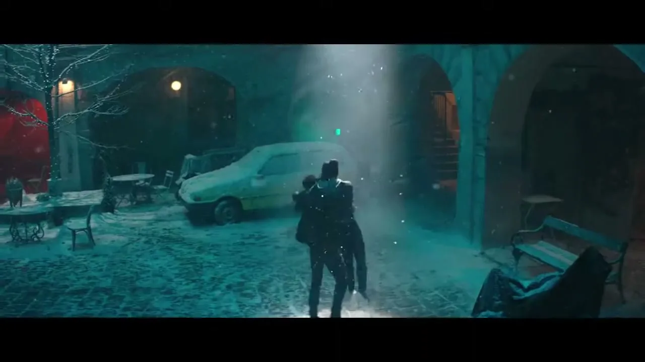A man and a woman dance in a snowy, dimly lit courtyard at night, with a car and tables in the background.