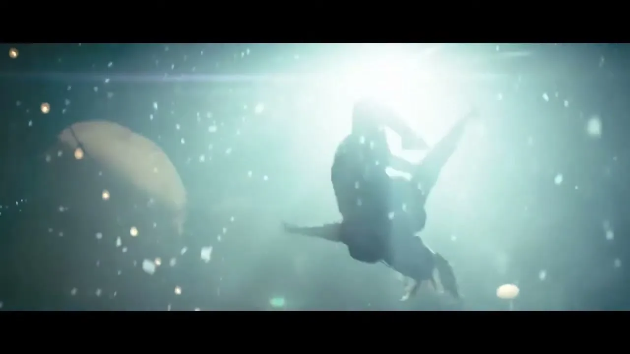 A male dancer in dark clothing performs an acrobatic move, silhouetted against a bright spotlight in a dark, misty space with floating particles.