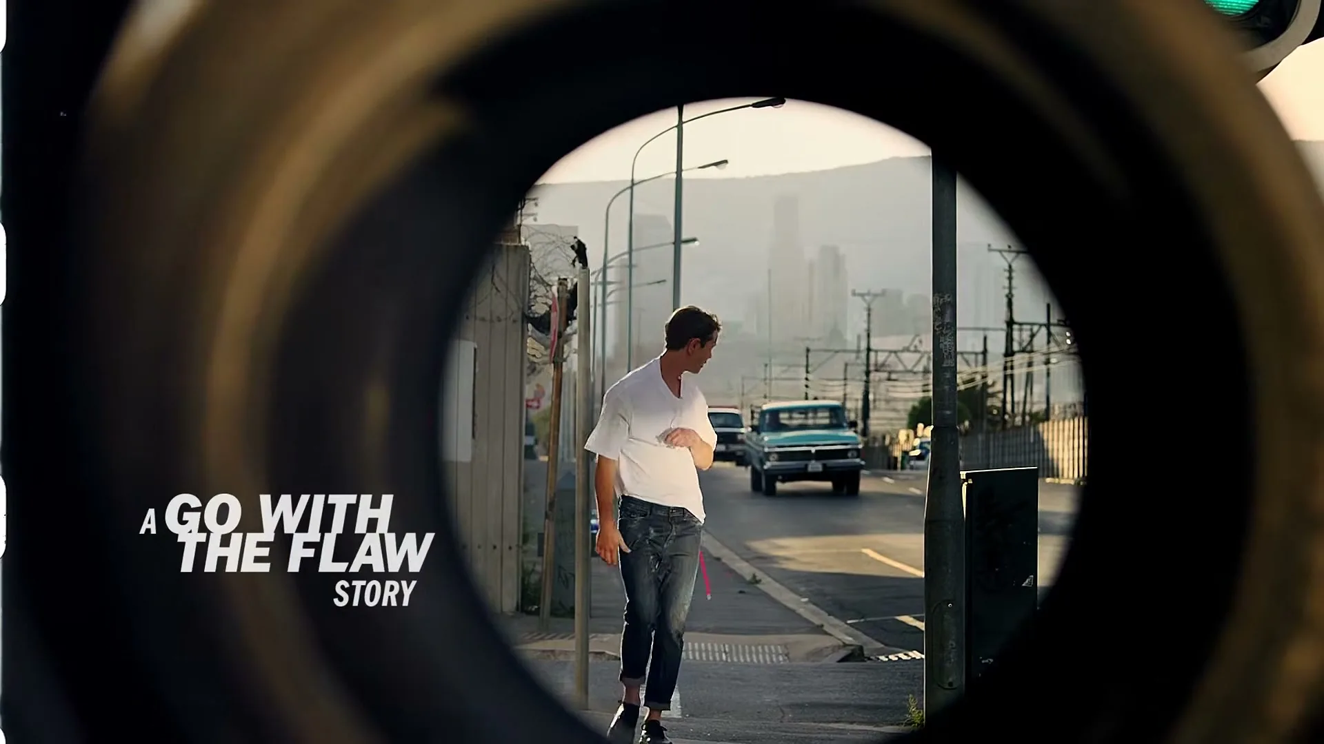 A young man in a white shirt and jeans walks down a street, framed through a circular opening, with a city and mountains in the background.