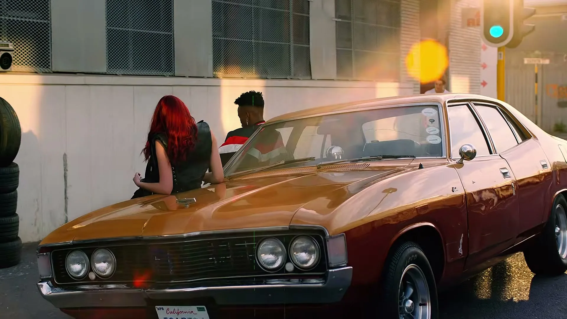 A young woman with red hair leans on the hood of a vintage car, while a young man sits in the driver's seat on a city street.